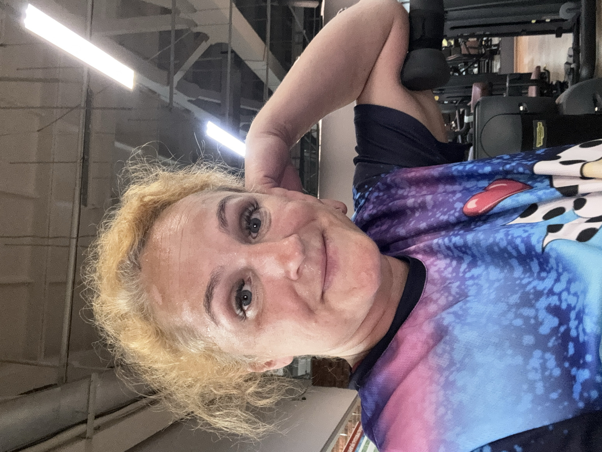 A selfie is taken in a gym, with the subject, a woman with blonde, curly hair and visible sweat on her forehead, looking towards the camera. She is wearing a t-shirt with a galaxy-like pattern in shades of blue, purple, and pink. Her left arm is bent and resting on her head, with a dark object, possibly a dumbbell, partially visible near her elbow. The background shows gym equipment, including weight machines and lighting fixtures on the ceiling, suggesting an indoor workout session. The overall lighting is bright, consistent with artificial gym lighting. The image's orientation is portrait, but the content is captured sideways. No visible text is present in the image. The context of Chaikovsky, Russia, places the scene geographically.