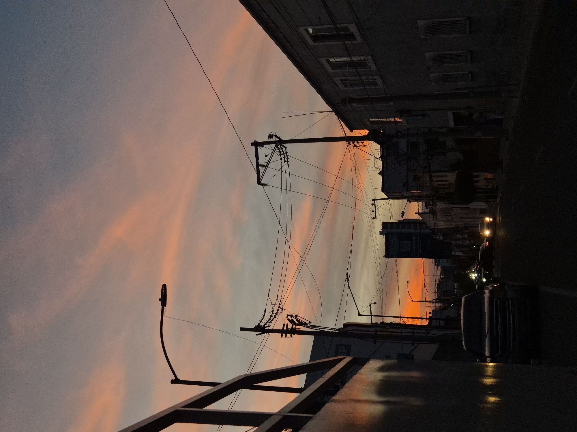 This media file captures an urban street scene during twilight, likely sunset, in Puebla, Mexico. The sky dominates the upper portion of the image, showcasing soft blends of pink and orange clouds transitioning into a cooler gray-blue.
Numerous utility poles and a complex web of overhead power lines crisscross the sky, creating a strong silhouette against the colorful backdrop. On both sides of the street, buildings rise, appearing as dark silhouettes with some details like windows and air conditioning units faintly visible, particularly on the building to the left.
The street below is dark, reflecting the low light conditions. Several vehicles are present, primarily visible as dark shapes with illuminated headlights and taillights, suggesting ongoing traffic. A prominent light-colored vehicle, possibly a van or sedan, is parked or moving in the right foreground. Towards the mid-ground on the right, a large billboard or building sign is partially visible, featuring text that appears to include words like "CONDOMINIOS," "LOFTS," and "VENTA" (Spanish for "sale"), along with a visible QR code, suggesting a real estate advertisement.
In the immediate foreground, a dark, angular structure, possibly part of a railing, building edge, or antenna, adds to the urban texture. The overall mood is calm and contemplative, highlighting the beauty of the twilight sky against the stark geometry of the city.