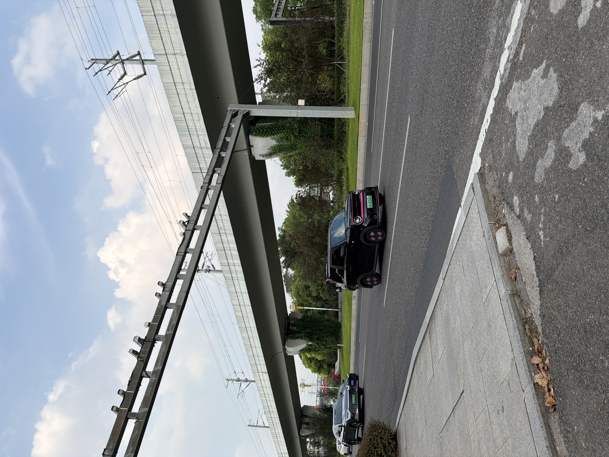 In Jiangjin, China, an overpass with overhead power lines runs horizontally across the frame, supported by concrete pillars partially covered in greenery. Below the overpass, a paved road accommodates vehicular traffic. In the foreground, a gray car is visible on the left, and a black electric microcar with pink accents on its wheels and bumper is driving on the right. The road is bordered by a concrete sidewalk and sparse vegetation. The sky above is partly cloudy.