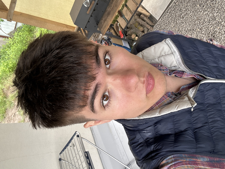 A young man with dark hair and a serious expression is wearing a dark blue vest over a red and black plaid shirt. He is standing outside, possibly in a backyard or patio area. The coordinates provided (latitude: 50.235, longitude: 30.386) are for Krushynka, Ukraine. The background of the image shows a patio area with a metal folding clothes rack, a grill, and a pile of wood. The surface below the man is covered in small, gray stones. There are additional tools and building materials visible to the right of the man. The scene is captured from a low angle, looking up at the man's face. The man appears to be taking a selfie.