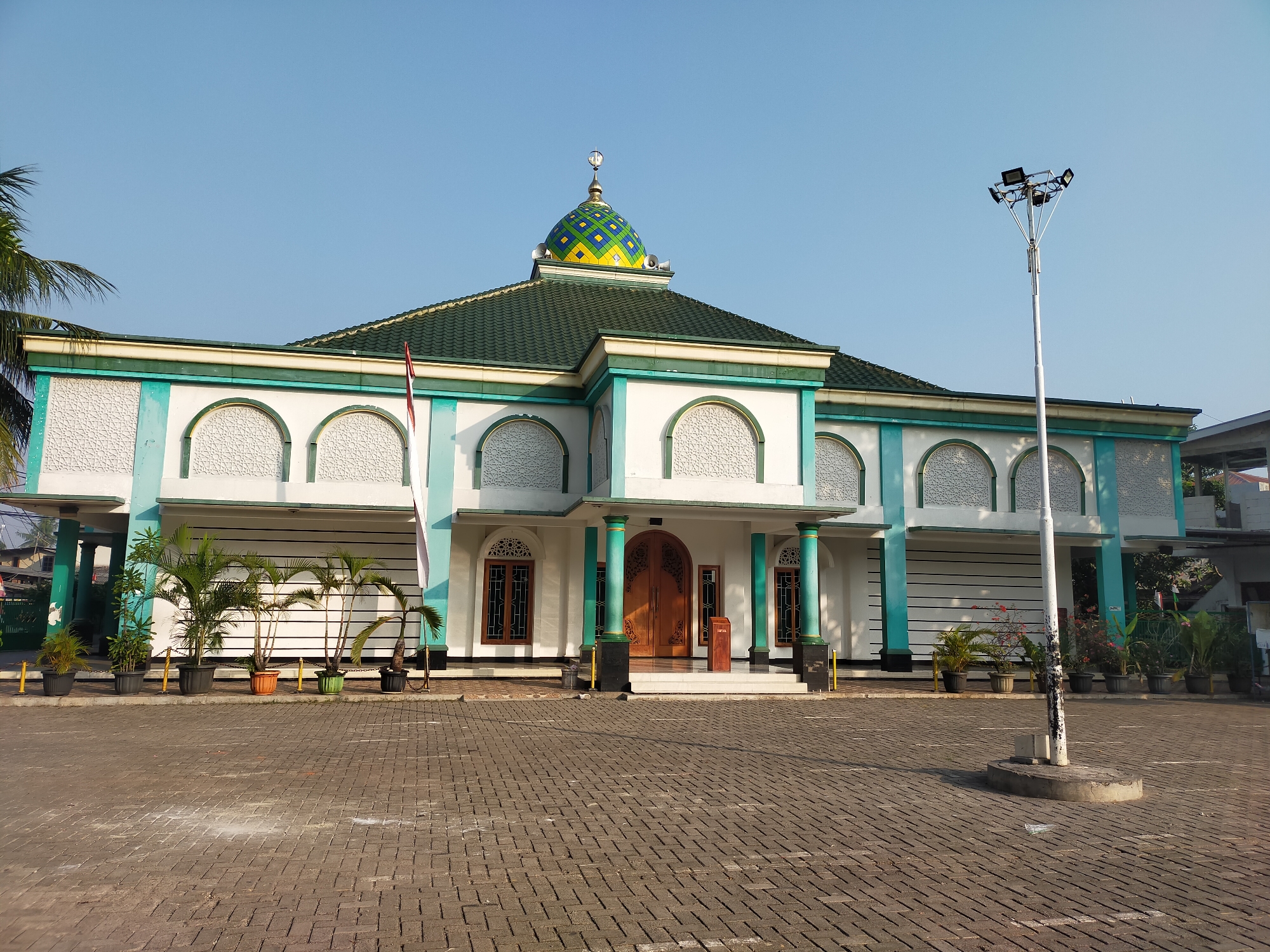 The image shows a mosque in Tambun, Indonesia, under a clear blue sky. The mosque features a green tiled roof, white walls with teal accents, and a colorful dome with yellow and blue geometric patterns. The entrance is flanked by wooden doors and windows with decorative grilles. Potted plants and palm trees are arranged in front of the building, and a paved courtyard with interlocking brick pavers extends outwards. A tall lamppost with multiple floodlights stands to the right of the mosque. The scene appears peaceful and is bathed in the soft light of either early morning or late afternoon. There are no people visible in the image.