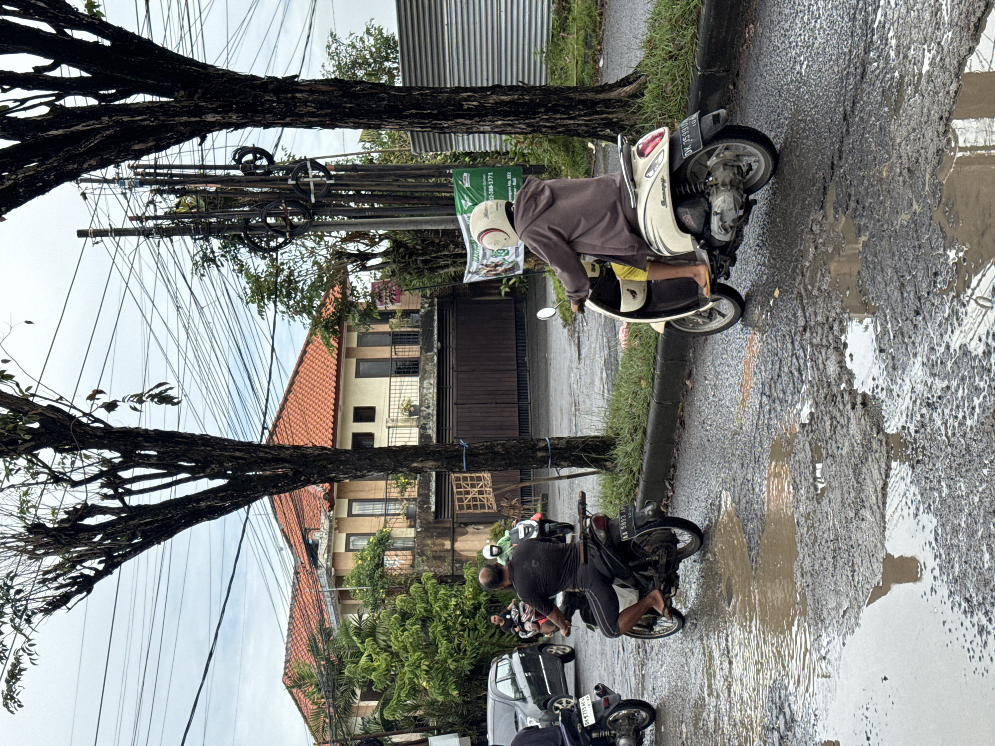 This media file captures an everyday street scene in Kuta Selatan, Indonesia, likely after a rain shower, given the numerous muddy puddles and extensive potholes marring the asphalt road. The sky is overcast, indicating daytime with soft, diffused light.
The foreground features a large, gnarled tree trunk on the left, partially obscuring a dense network of overhead utility wires strung between poles. The road surface is severely damaged, filled with standing water and reflections of the sky and surroundings.
Several individuals are visible, primarily on motorcycles. A person wearing a brown hoodie and a white helmet is paused on a cream-colored Honda Scoopy scooter, positioned on the left side of the road. Its license plate, "DK 3590 V", is partially visible. Further down the street, another person, bald and dressed in a black t-shirt and shorts, is on a dark motorcycle (license plate "AA 5893 VY"), also facing away from the camera. Other motorcycles and a dark car are partially visible further along the street, navigating the challenging road conditions.
In the background, buildings with distinctive orange-tiled roofs and balconies are visible, typical of Indonesian architecture. A green banner hangs from a utility pole near the cream scooter, displaying text including "Indonesian Driver," "02-XXX-1771," "Denpasar No. 222," and "Kerobokan Kelod," providing clear location cues. The scene depicts a common street environment in Indonesia, highlighting the reliance on two-wheeled transport and the challenges posed by infrastructure.