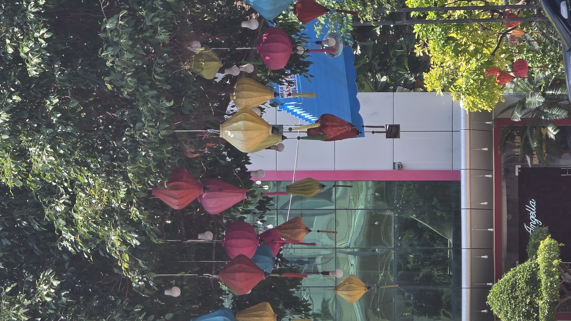The image depicts a street scene in Nha Trang, Vietnam. Colorful paper lanterns hang suspended above the street, intermingling with lush green foliage. A modern building with a mirrored facade and pink trim is visible behind the lanterns. A sign reading "Angella" is displayed on the building. A blue awning is also visible.