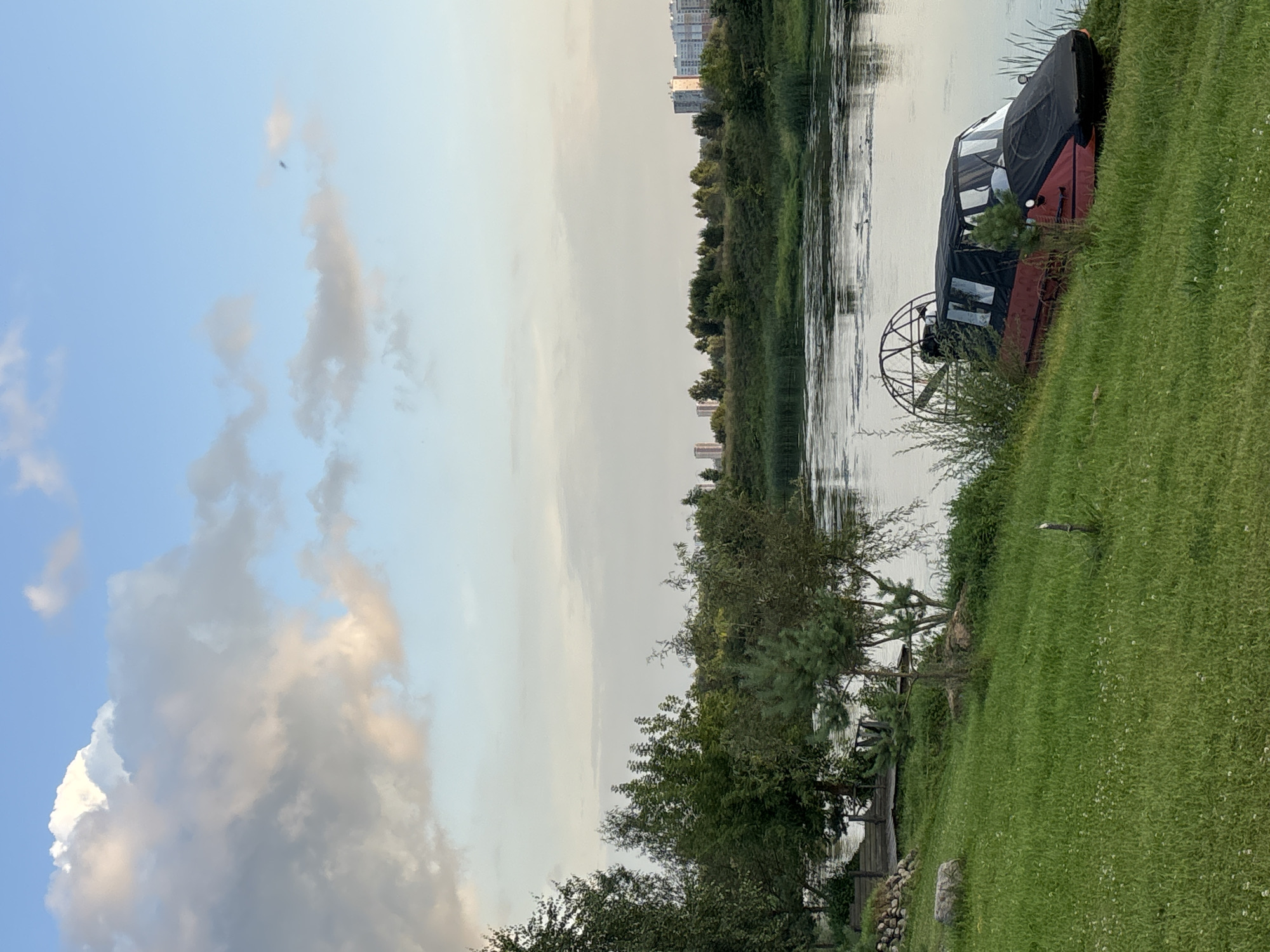 An airboat with a red hull and a black canopy is parked on a grassy incline next to a body of water. The airboat has a large fan mounted at the rear. The water is calm, reflecting the sky, and has some surface debris. The grassy bank is covered in green grass and small white flowers, with a few scattered rocks and a small wooden post. In the background, there are lush green trees and a line of buildings in the distance, suggesting an urban or suburban environment. The sky is partly cloudy with large, soft clouds in shades of white and gray against a light blue sky. This scene is located in Zvenigorod, Russia.