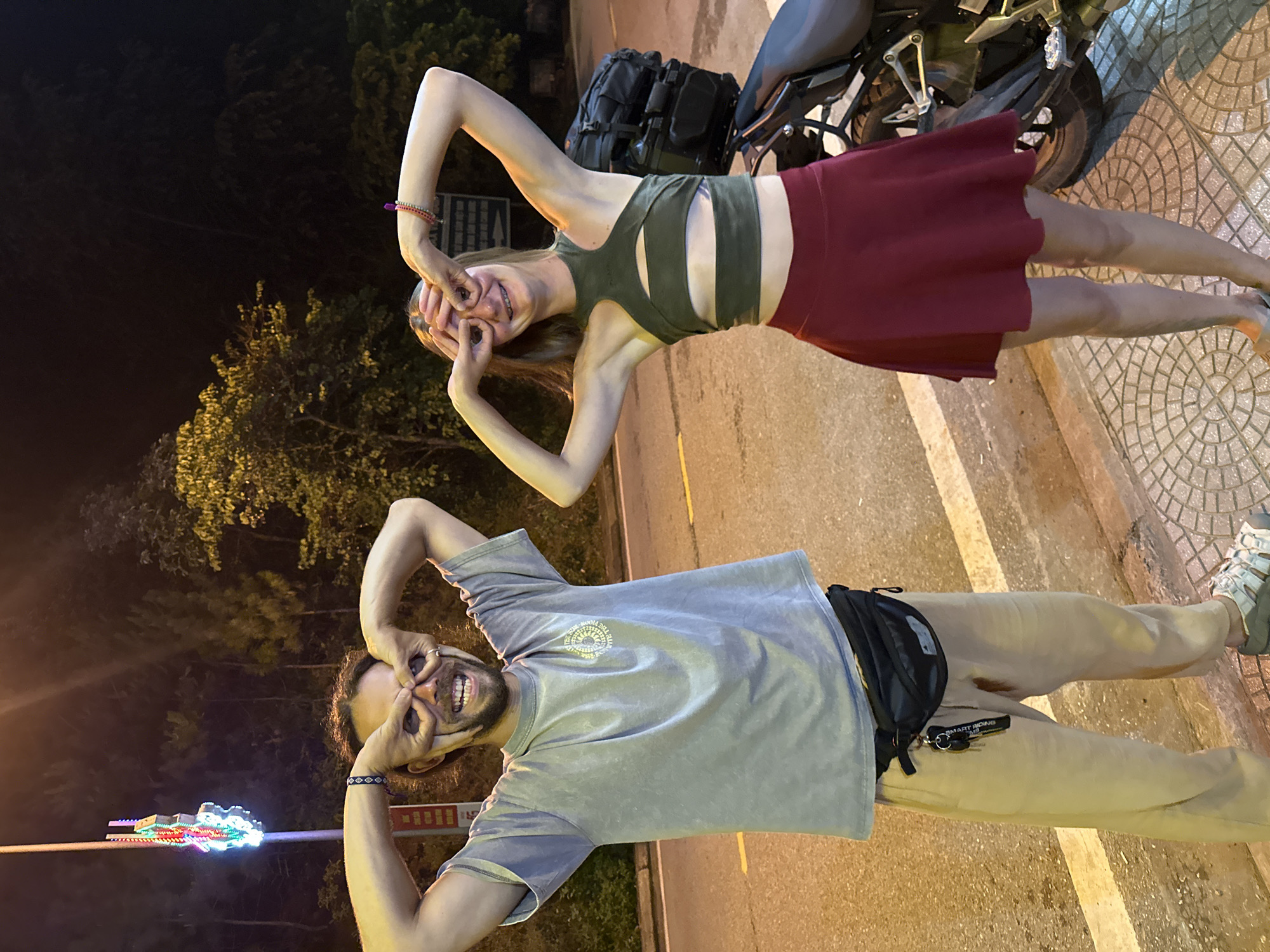 A nighttime outdoor photograph captures a man and a woman posing playfully, illuminated by artificial light against a dark background of trees, likely in Mù Cang Chải, Vietnam. The image is rich with expressions of joy and casual travel details.
On the right, a woman with light brown hair smiles brightly, her hands cupped around her eyes in a playful "binoculars" gesture. She wears a dark olive green crop top with cutouts and a maroon skirt, accented by a colorful beaded bracelet on her left wrist. She stands on a patterned tiled sidewalk, beside a black motorcycle equipped with a black backpack.
On the left, a man with dark hair, a beard, and a mustache mirrors her pose, grinning widely with his hands similarly cupped around his eyes. He is dressed in a light gray t-shirt featuring a subtle circular graphic (partially readable as "THE FLOOR IS...") and light brown pants. A black fanny pack, from which a keychain with "SMART RIDING M3" hangs, is fastened around his waist. He also wears a braided bracelet and a ring. Near him, a tall pole adorned with bright, colorful LED lights adds a festive touch to the scene.
Both individuals exude happiness and playfulness, suggesting a lighthearted moment during an evening outing or journey. The setting includes a paved road with yellow markings and trees in the distance, characteristic of a roadside stop in a scenic area.