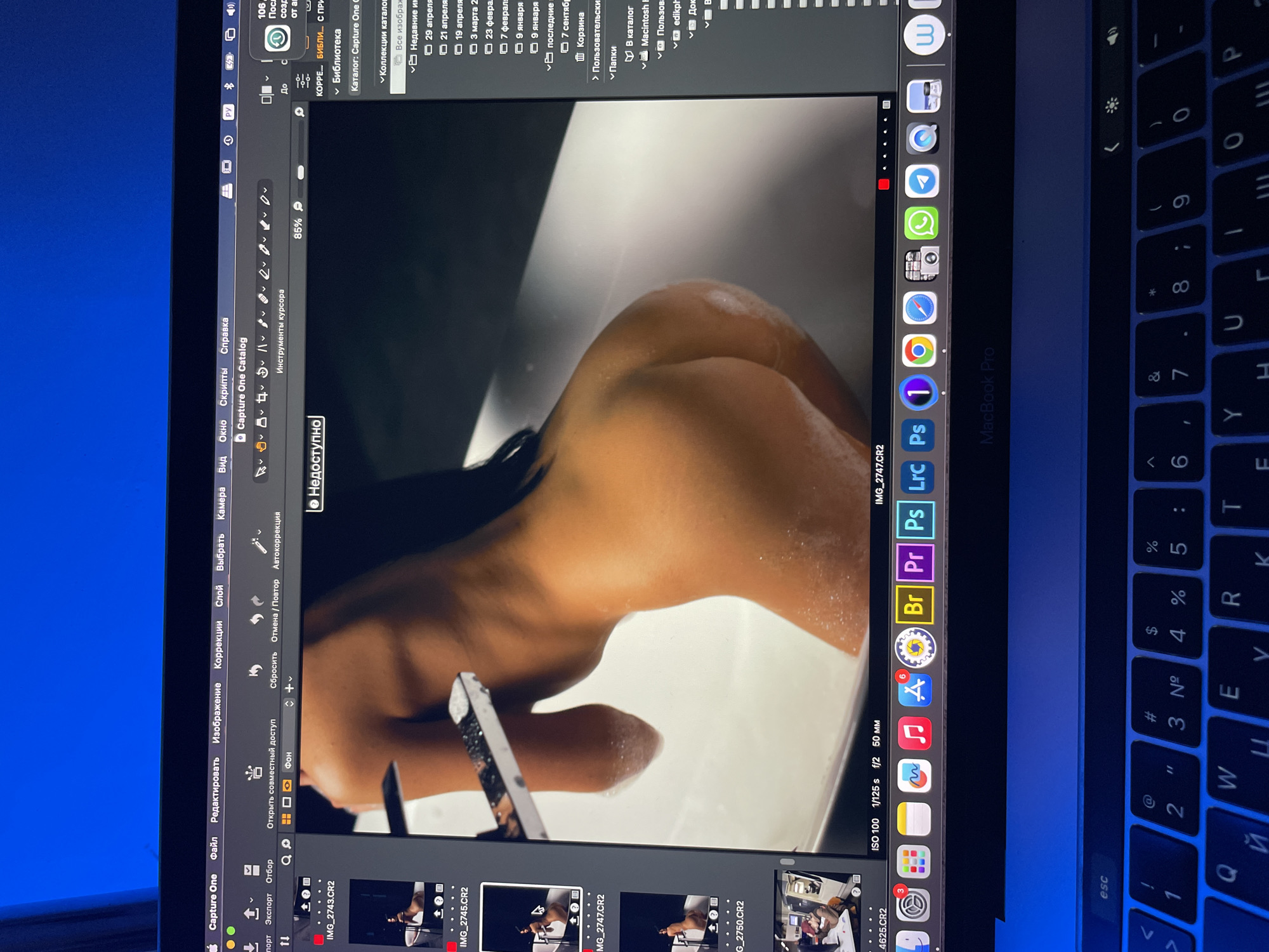 The image is of a computer screen displaying an image of a nude woman's back and shoulder in Capture One photo editing software. The screen is likely part of a Macbook Pro, with the keyboard visible. Other icons are visible on the dock, including Google Chrome, WhatsApp, and Finder. The software is in Russian. ISO 100, 1/125 s, f/2, 50 mm is displayed on the software interface. The location is not visible.