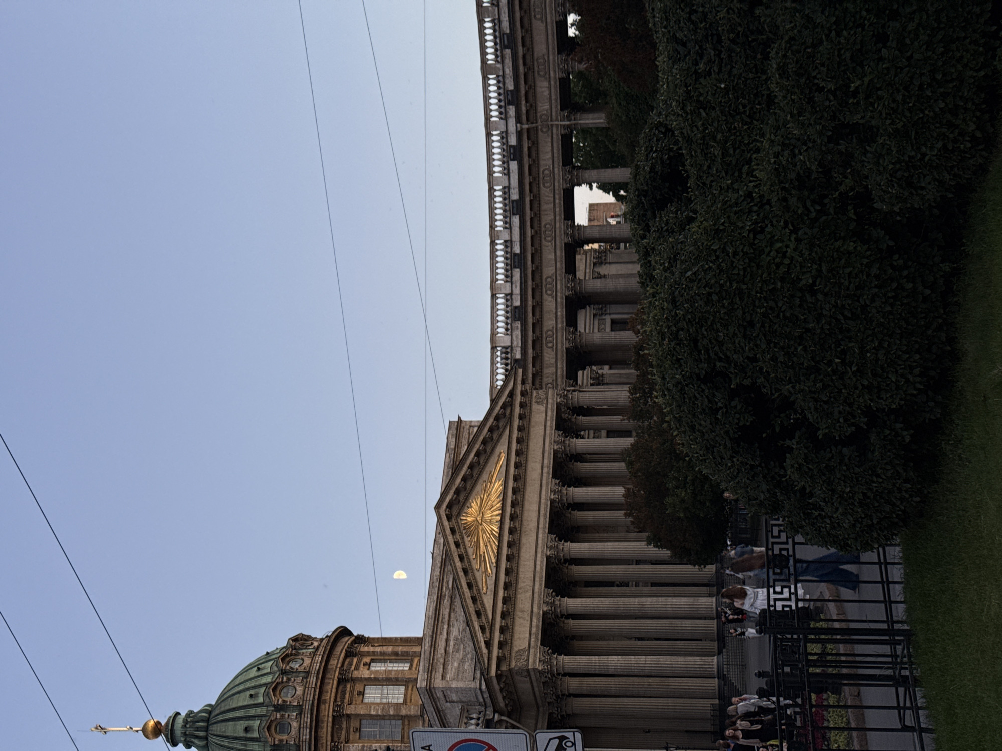 The image shows the Kazan Cathedral in Saint Petersburg, Russia. The cathedral is a prominent landmark, known for its neoclassical architecture and its impressive semicircular colonnade. In the foreground, there is a black metal fence with a pathway behind it, and then the colonnade and part of the dome of the cathedral. Behind the fence, several people are visible, some walking and others standing. The sky is clear and a pale blue, with a half-moon visible. The overall impression is of a late afternoon or early evening scene, with the soft light casting long shadows.