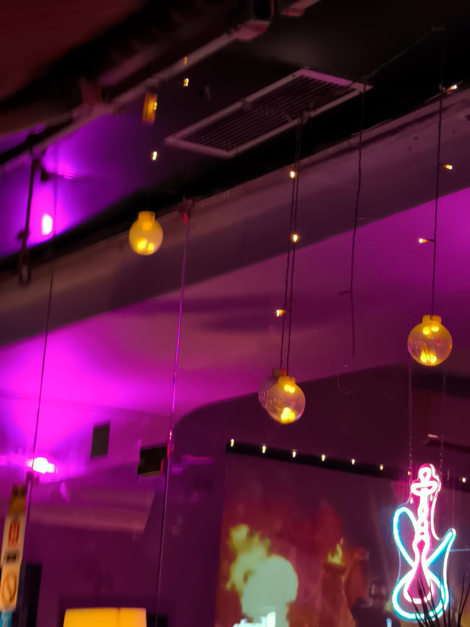 The image is an interior shot of a dimly lit establishment, possibly a bar or lounge, with a strong purple ambient lighting. Several strings of small, round, illuminated yellow lanterns hang from the ceiling. In the background, a neon sign shaped like a hookah is visible on the right side. The environment appears to be decorated with a mix of modern and decorative elements, including what looks like a ventilation grate on the ceiling. The overall impression is one of a vibrant, atmospheric nightlife venue. This scene is located at "Boom Beach Batumi," a popular nightclub and lounge situated on the Batumi Boulevard in Batumi, Georgia.