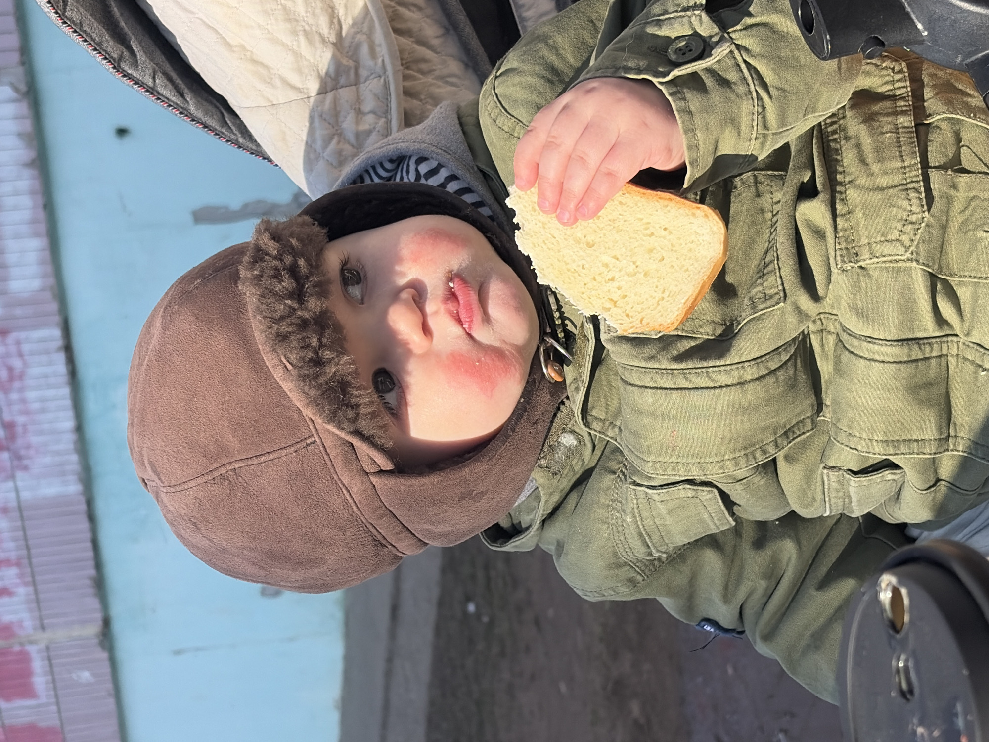 This image depicts an outdoor scene in the city of Chaikovsky, Russia, focusing on a young child, likely a toddler, seated and actively holding a slice of bread. The child wears a brown ear-flapped hat with visible faux fur lining and a green, multi-pocketed military-style jacket, under which a grey hooded garment with a black and white striped inner pattern is visible. The child's cheeks are notably red, and bread crumbs adhere to the lower lip as the gaze is directed slightly upward and to the right. The right hand firmly grasps a thick, light-colored slice of bread. The child is positioned in what appears to be a stroller or similar seating apparatus, evidenced by a quilted, cream-colored material behind the back and a dark structural component in the lower left foreground. The immediate background features a light aqua-blue wall or fence, exhibiting surface wear and a faded pink section along its upper edge, indicative of a corrugated texture. Below the wall, a patch of brown, dry ground is visible. The scene is brightly illuminated by direct sunlight.