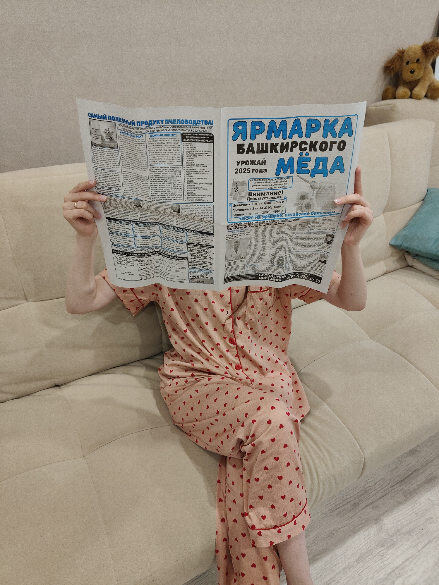 A person, whose face is obscured by a large open newspaper, is sitting on a light beige upholstered couch. The individual is wearing light pink pajamas with a red heart pattern. Their hands, with a ring visible on the left ring finger, hold the newspaper open.
The newspaper, written in Russian, prominently features an advertisement on the right side for a "Bashkir Honey Fair" (ЯРМАРКА БАШКИРСКОГО МЁДА) for the "Harvest 2025" (УРОЖАЙ 2025 года). It announces a "Special offer!" (Внимание! Действует акция!) for various types of honey, including floral, buckwheat, and mountain honey, with discounted prices (e.g., 3 kg of floral honey for 1390 rubles). The advertisement also mentions "Altai balms" (алтайские бальзамы), invites readers to the fair with listed dates and times, and advertises "Free Delivery" (БЕСПЛАТНАЯ ДОСТАВКА) along with a phone number (8 (912) 878-26-50).
On the left side, an article is titled "The Most Useful Beekeeping Product!" (САМЫЙ ПОЛЕЗНЫЙ ПРОДУКТ ПЧЕЛОВОДСТВА!), discussing "Royal Jelly" (МАТОЧНОЕ МОЛОЧКО). The text highlights its ability to boost immunity and lists various ailments for which it is recommended, such as atherosclerosis, post-surgery recovery, arthritis, cataracts, and vision weakness. Sections titled "Interesting Fact" (ИНТЕРЕСНЫЙ ФАКТ) and "Important Moment" (ВАЖНЫЙ МОМЕНТ) are also visible.
The scene is an indoor setting, likely a living room. A light blue throw pillow rests on the couch to the right, and a brown, shaggy plush dog toy is visible on the backrest of the couch in the upper right corner of the frame. The lighting suggests a well-lit environment.
