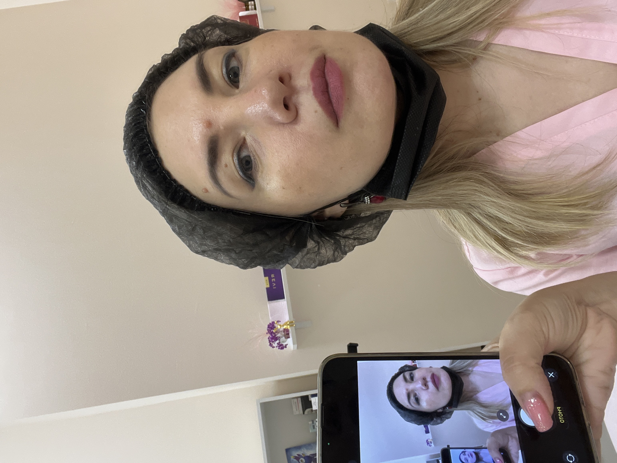 The image shows a close-up of a woman's face, with a black hairnet and a black mask covering parts of her face. She is wearing a pink robe. Her lips are a dark pink color, and she has noticeable eye makeup. There is a red mark or blemish on her cheek. The background is a lightly colored wall. A smartphone is held in the foreground, taking a picture of the woman. The phone's screen shows a reflection of the woman, also wearing the hairnet and mask, and the pink robe. The phone has a camera interface visible, with a "photo" button and other icons. The overall lighting suggests an indoor setting, possibly a beauty salon or spa, given the hairnet and the woman's makeup. The context provided indicates the location is Sochi, Russia. There is no indication of the time of day or weather. The woman's expression is neutral. Some blurred text is visible on a purple box on a shelf in the background, but it is unreadable.