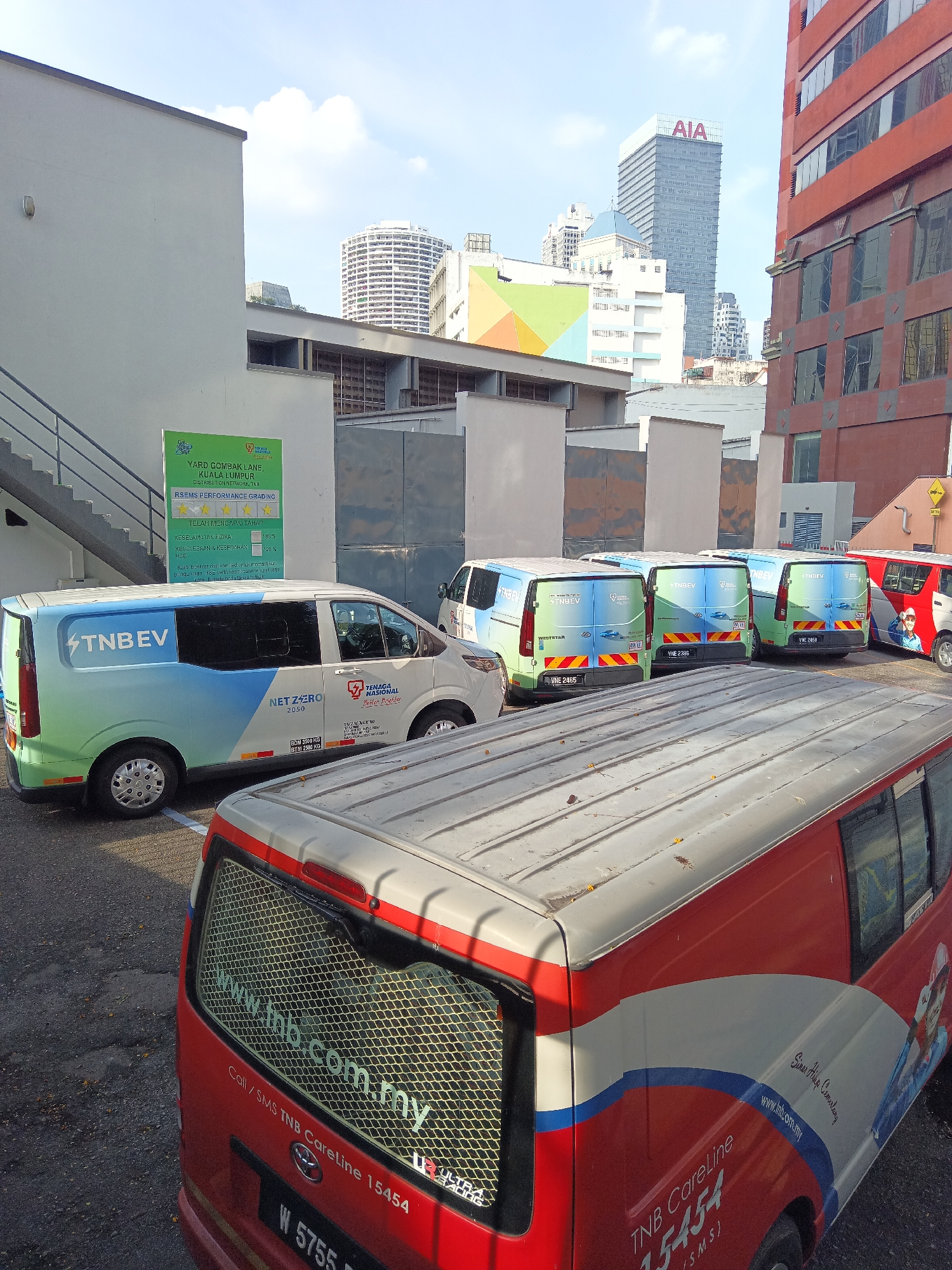 The image shows a parking lot in Kuala Lumpur, Malaysia, filled with several vans, some of which are branded with "TNB EV" and "TNB CareLine". In the background, there are tall buildings, including one with the "AIA" logo. The environment appears to be an urban setting, possibly a depot or a service area for the utility company Tenaga Nasional Berhad (TNB). The vans are parked closely together, suggesting a working area. The sky is partly cloudy.