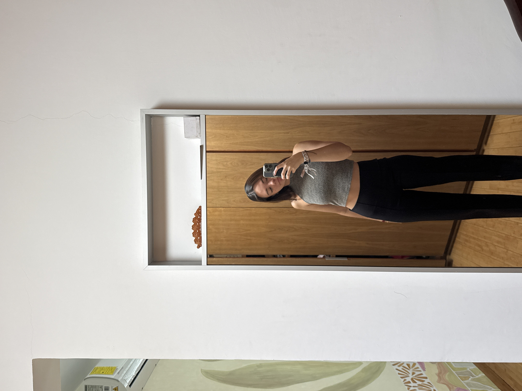 A woman is taking a selfie in a mirror, wearing a gray top and black leggings. The mirror reflects a room with wooden cabinets and flooring. The room appears to be located in a building with white walls. An air conditioning unit is visible in the lower left corner of the image. Based on the information provided, it is not possible to definitively confirm the location as San Bernardino Tlaxcalancingo, Mexico.