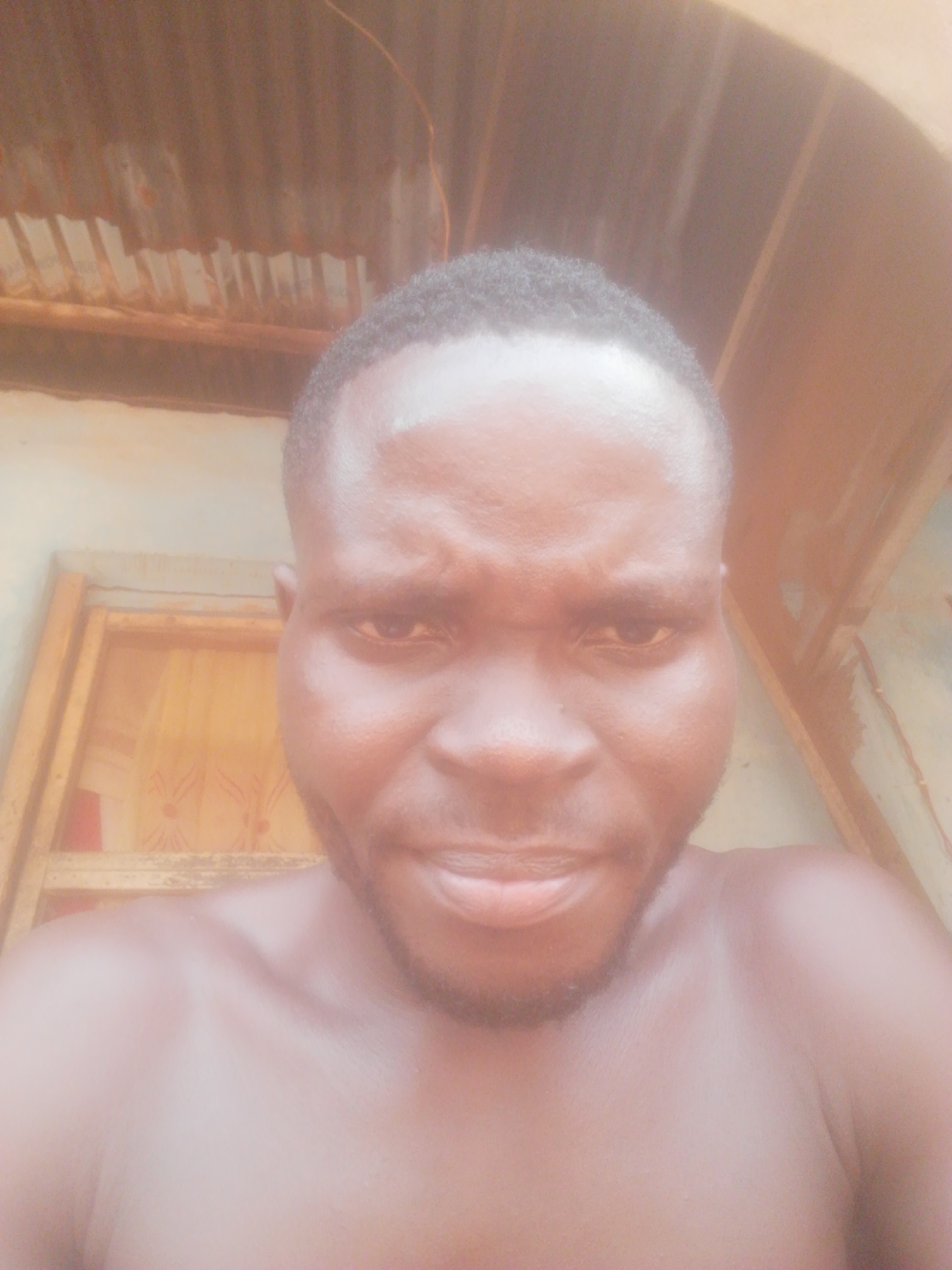 A headshot of a dark-skinned man with short, dark hair and a trimmed beard is centered in the frame. He appears to be shirtless and is looking directly at the camera with a somewhat serious expression. The background is slightly out of focus, but it suggests an outdoor setting. A corrugated metal roof is visible at the top, and a partially visible door or window frame is on the left. The location is unidentified.