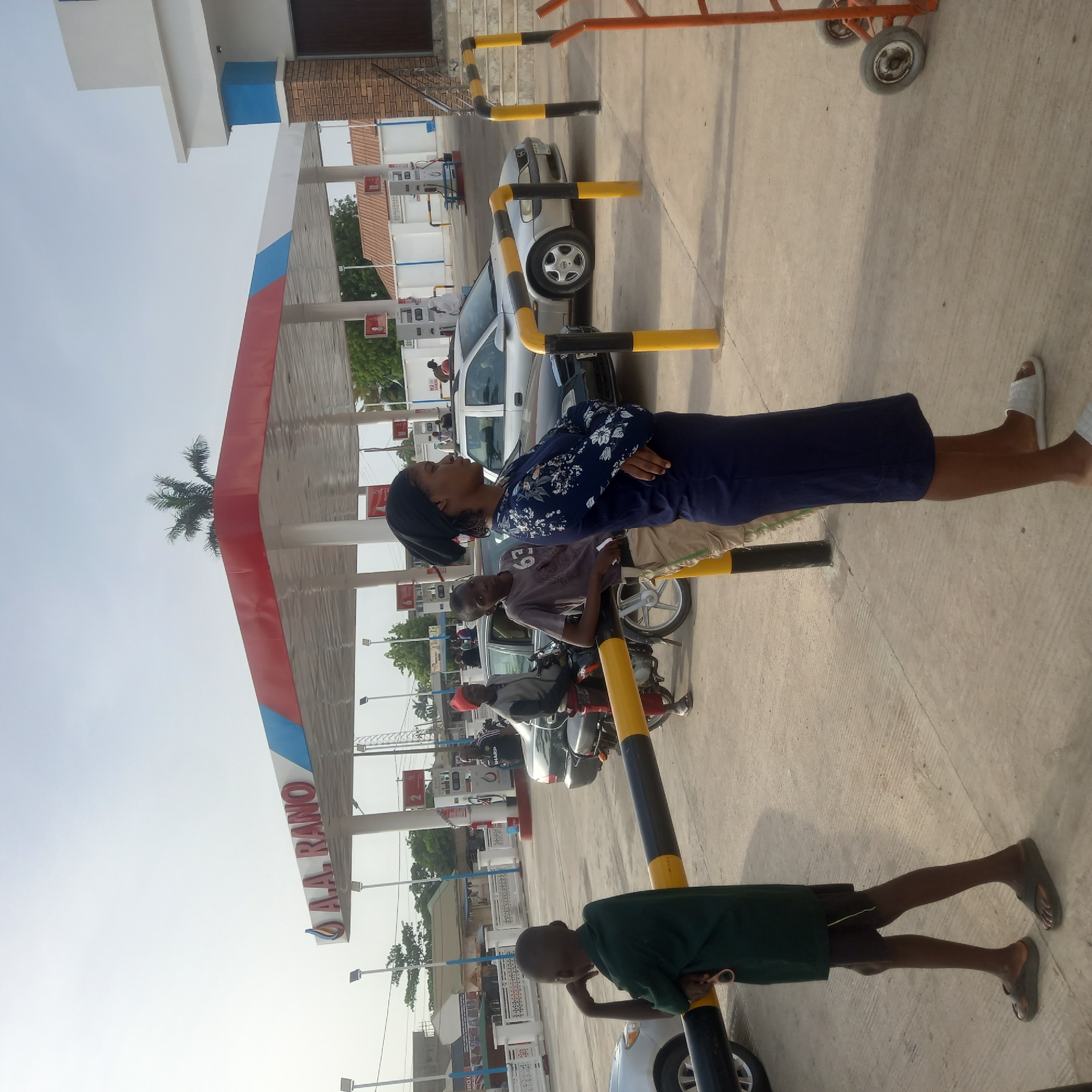 The image shows the outside of a AAA Rano gas station in what is presumed to be the city of Barden Yana, Nigeria. There is a gas station canopy with pumps underneath. A woman wearing a dark blue floral blouse and a headscarf is standing with her arms crossed. A young man is leaning against a yellow and black bollard. Several vehicles including cars and motorbikes are parked. A younger boy in a green shirt is standing by another bollard.