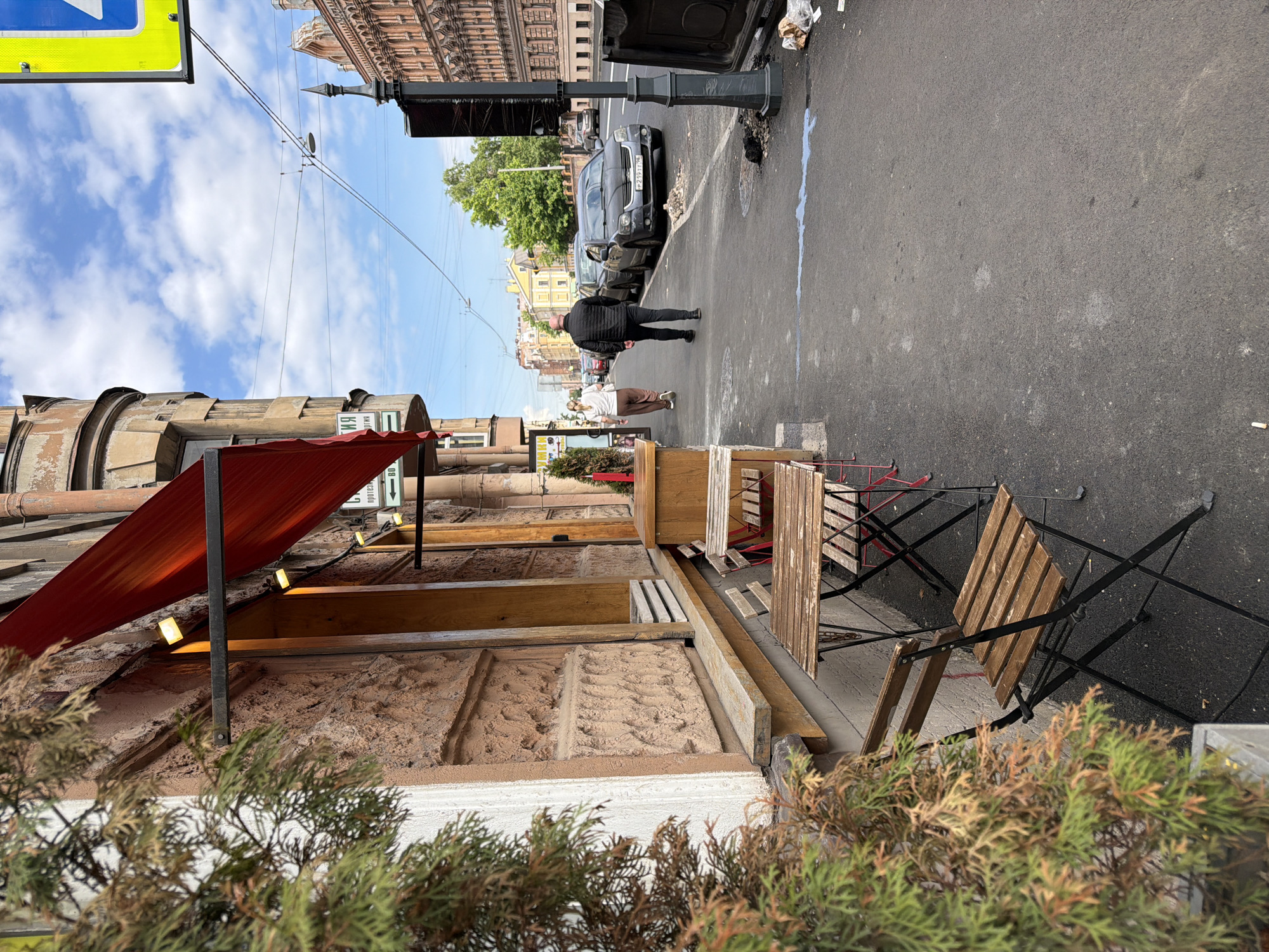 The image captures a street scene in Saint Petersburg, Russia, from a slightly elevated perspective, looking down a sidewalk and road. The sky is partly cloudy and bright, indicating daytime.
In the foreground on the right, there are outdoor cafe tables and chairs, some folded and stacked, suggesting the cafe might be closed or preparing to open/close. Planters with evergreen shrubs are visible along the building's edge. A red awning extends from the building, covering part of the sidewalk seating area, with small lights underneath. The building facade in the foreground is rough, textured stone or plaster.
Two individuals are seen walking away from the camera on the asphalt sidewalk towards the background. One person wears a black jacket and dark pants, and the other wears a white top and brown pants. Beyond them, a dark-colored SUV is parked on the street, displaying a Russian license plate "P 211 TT 177". Another dark sedan is parked behind it.
The street is asphalt, with a faint blue/white mark or spill running along the curb. Overhead electrical wires crisscross the sky, typical for tram or trolleybus routes in Russian cities. Buildings with classical European architecture line the street in the mid-ground and background, featuring ornate details. A large blue and yellow traffic sign is partially visible in the upper left, showing a white arrow. Near the cafe awning, vertical signs display partial Russian text fragments, including "ВИ" and "ВО", and another showing "до" and "60". A dark street lamp post stands near the parked cars. The overall scene depicts a typical urban environment.