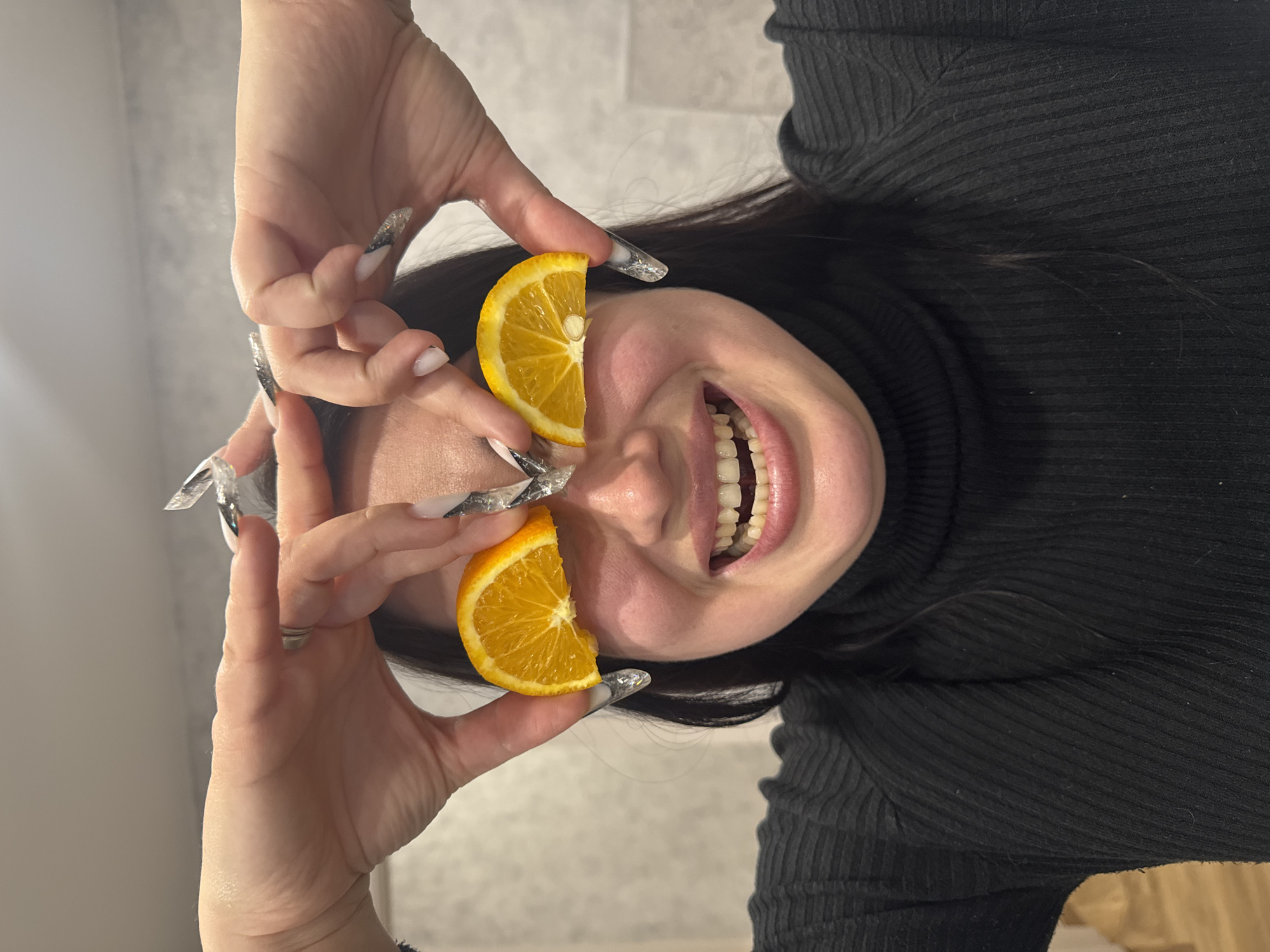 An indoor, eye-level shot captures a person playfully covering their eyes with two vibrant orange slices while smiling broadly. The subject, likely a woman, has long dark hair and is wearing a black ribbed turtleneck top. Her hands are positioned on either side of her face, with fingers holding the orange slices over where her eyes would be.
Her mouth is open in a wide, joyful smile, revealing her teeth. The hands feature long, decorative acrylic nails, which appear to be clear or silver with intricate designs, possibly glitter or foil. One orange slice clearly shows a small seed. The background is a soft, light-colored wall with a subtle texture, out of focus, suggesting an indoor setting. The overall mood of the image is lighthearted and cheerful. There is no visible text in the image.