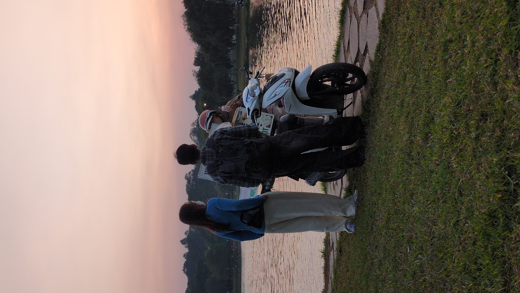 A scene captured in Da Lat, Vietnam, shows three individuals positioned alongside a body of water with a white scooter parked nearby. The lighting suggests either sunrise or sunset. One person, facing the water, wears a blue top and light-colored pants. Another individual, dressed in a plaid shirt and dark pants, stands next to the scooter, appearing to observe the scene. On the scooter, a person wearing a helmet seems to be eating something. The scooter has a sign attached to the back. The background includes a body of water and trees.