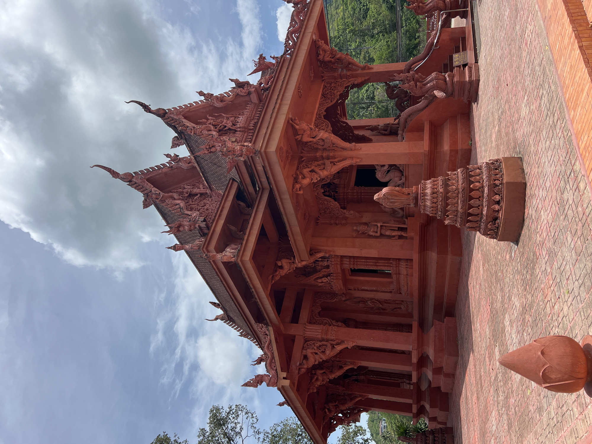 The image depicts a vibrant red temple structure situated in Baan Hua Thanon, Thailand. The temple features intricate architectural details, including multi-tiered roofs with ornate carvings and decorative finials. Statues of mythical creatures and figures are integrated into the design, adorning the columns, eaves, and surrounding plaza. The plaza itself is paved with red bricks, and in the foreground, there are decorative lotus-shaped sculptures. Lush green trees and a hilly landscape are visible in the background, suggesting a tropical environment. The sky is partly cloudy, with a mix of blue patches and grey clouds. No people are visible in the image.