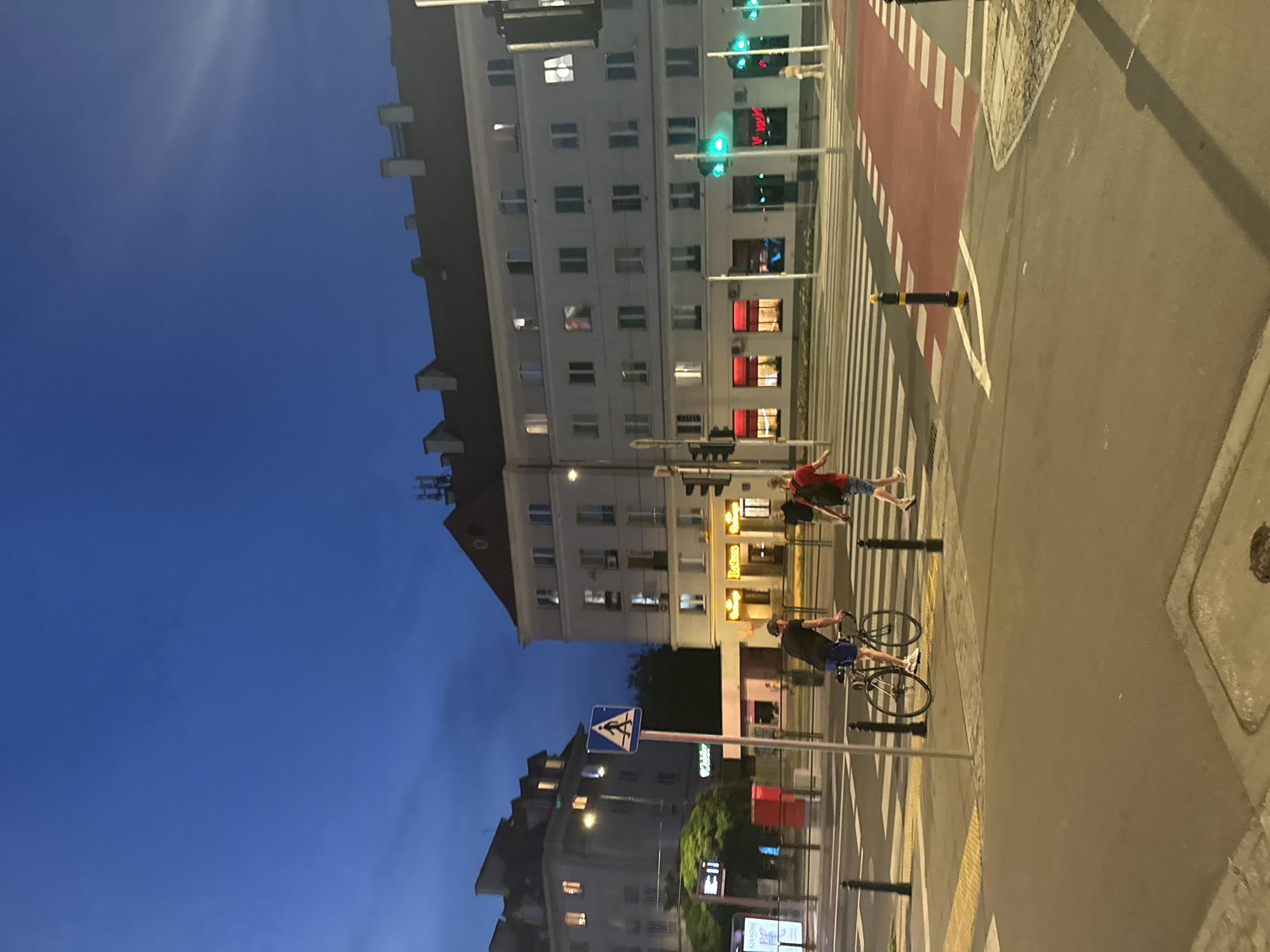 An evening shot captures a street scene at a crosswalk in an urban environment. The sky is a deep blue, indicating twilight. On the left, a person on a bicycle, wearing a dark t-shirt and blue shorts, is on the crosswalk, facing away from the camera. They are wearing a cap. To the right, two people are walking across the crosswalk. The person closer to the camera is wearing a red t-shirt and blue shorts, carrying a backpack. The other person, further away, is wearing a light-colored outfit.
Several buildings line the street. The building on the right is a multi-story structure with numerous lit windows. Red-lit storefronts are visible at street level, with signs that are difficult to decipher. Traffic lights are positioned at the intersection, with green lights illuminated. A pedestrian crossing signal with a walking figure is visible on a pole.
In the foreground, the crosswalk has alternating white and red striped sections. Black bollards are placed along the edge of the street. A street sign with a walking figure symbol is present. The street surface appears wet in some areas, reflecting the ambient light. The overall impression is of a busy intersection at dusk.