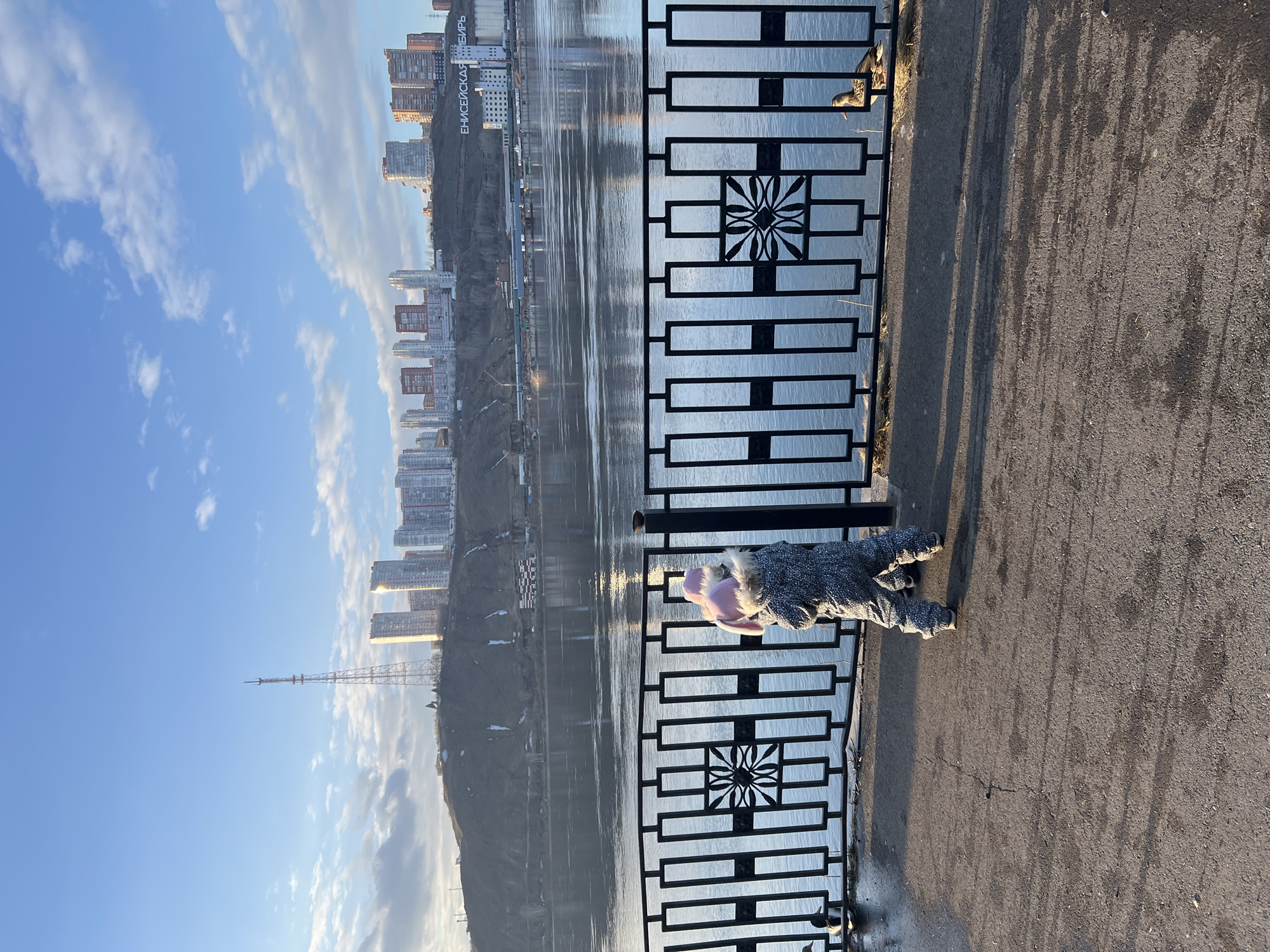 The image depicts a panoramic view of the Yenisei River embankment in Krasnoyarsk, Russia, likely during late afternoon or early morning dueolight and shadow. In the immediate foreground, a dark asphalt path shows long shadows cast by a black metal railing. Two young children, viewed from behind, stand facing the railing, looking towards the river. The child on the right wears a speckled grey snowsuit with a pink hat featuring ear-like protrusions. The child on the left is partially visible, also in a dark speckled snowsuit. The ornate black metal railing features repeating rectangular panels and a central decorative floral-like motif. Two ducks are visible on the water's edge near the railing: one on the far left and another on the far right. The wide, calm Yenisei River occupies the midground, reflecting the sky and distant buildings. On the opposite bank, a steep, dark hillside rises, partially covered by patches of snow or pale earth. At the base of this hillside, a large white building displays the Russian inscription "ЕНИСЕЙСКАЯ СИБИРЬ" (Yeniseyskaya Siberia). The background reveals the Krasnoyarsk city skyline atop the hillside, dominated by numerous multi-story residential buildings, some brightly lit by the low sun. A tall, slender lattice-structure television or radio tower stands prominently on the far left of the distant ridge. The sky above is clear blue with scattered white cumulus clouds.
