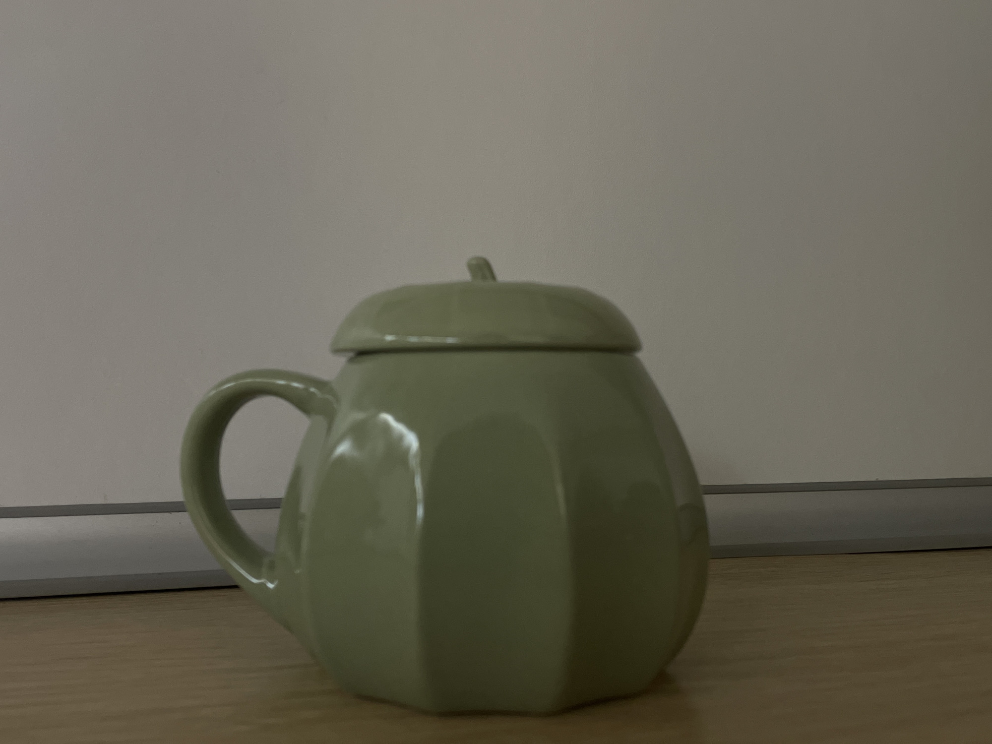 A light green mug with a lid sits on a wooden surface. The mug is shaped like a pumpkin, with visible vertical indentations around its body. The lid also has a similar ribbed texture and a small stem-like knob on top. A handle is visible on the left side of the mug. The background is a plain white surface with a thin grey horizontal line near the bottom. The lighting appears to be from the front and slightly above, casting a subtle shadow to the right of the mug. The scene suggests an indoor setting, possibly a desk or table. No people, text, or specific time or weather cues are present.