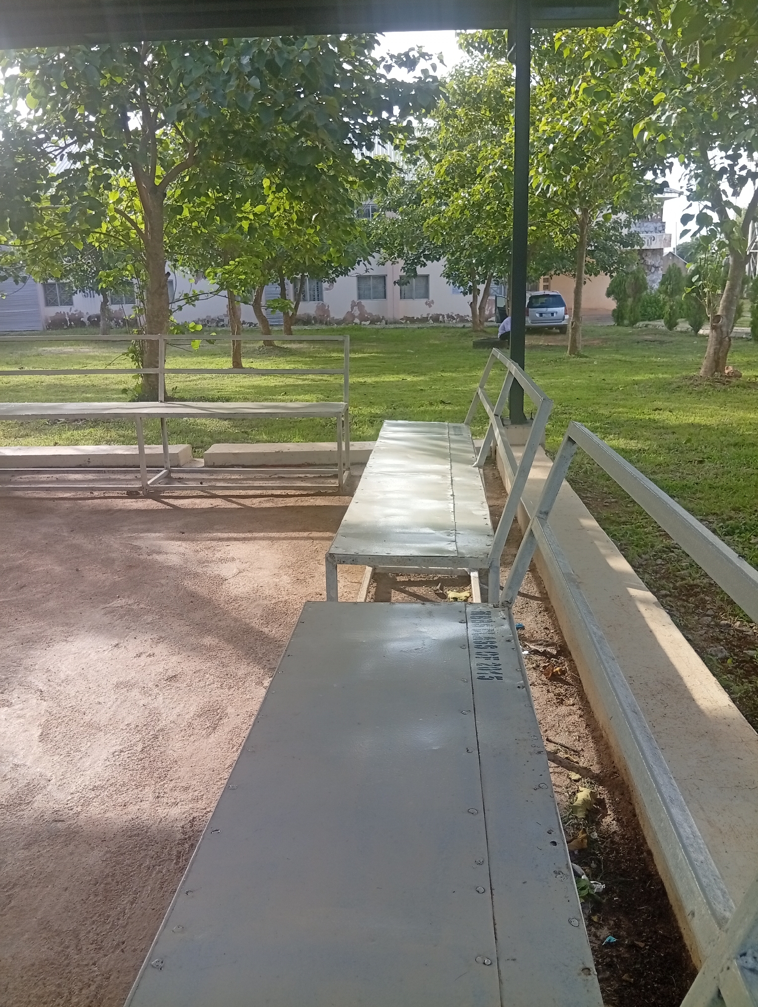 The image depicts an outdoor scene on a sunny day, likely in the late afternoon or early morning given the long shadows. The setting appears to be a park or a campus area, characterized by manicured green grass, trees with lush foliage, and a building in the background. There are several metal benches with backrests, suggesting a place for rest or waiting. The ground in the foreground is a light-colored dirt or sand, and a concrete structure runs alongside the benches.
The image focuses on the benches and the surrounding environment. One bench in the foreground is particularly detailed, showing its metallic construction and some visible rivets. On the side of this bench, faint text can be partially read: "2013". This suggests the bench might be dated or have some form of identification from that year.
In the background, a silver minivan is parked near the building, with its driver's side door open. A person, likely the driver, is partially visible sitting in the car. The building in the background is a light-colored, multi-story structure with windows. The overall atmosphere is tranquil and somewhat deserted, with the natural elements of trees and grass dominating the scene. The sunlight creates a dappled effect on the ground and benches, highlighting the textures of the environment. No specific event or activity is clearly taking place, implying a moment of stillness.