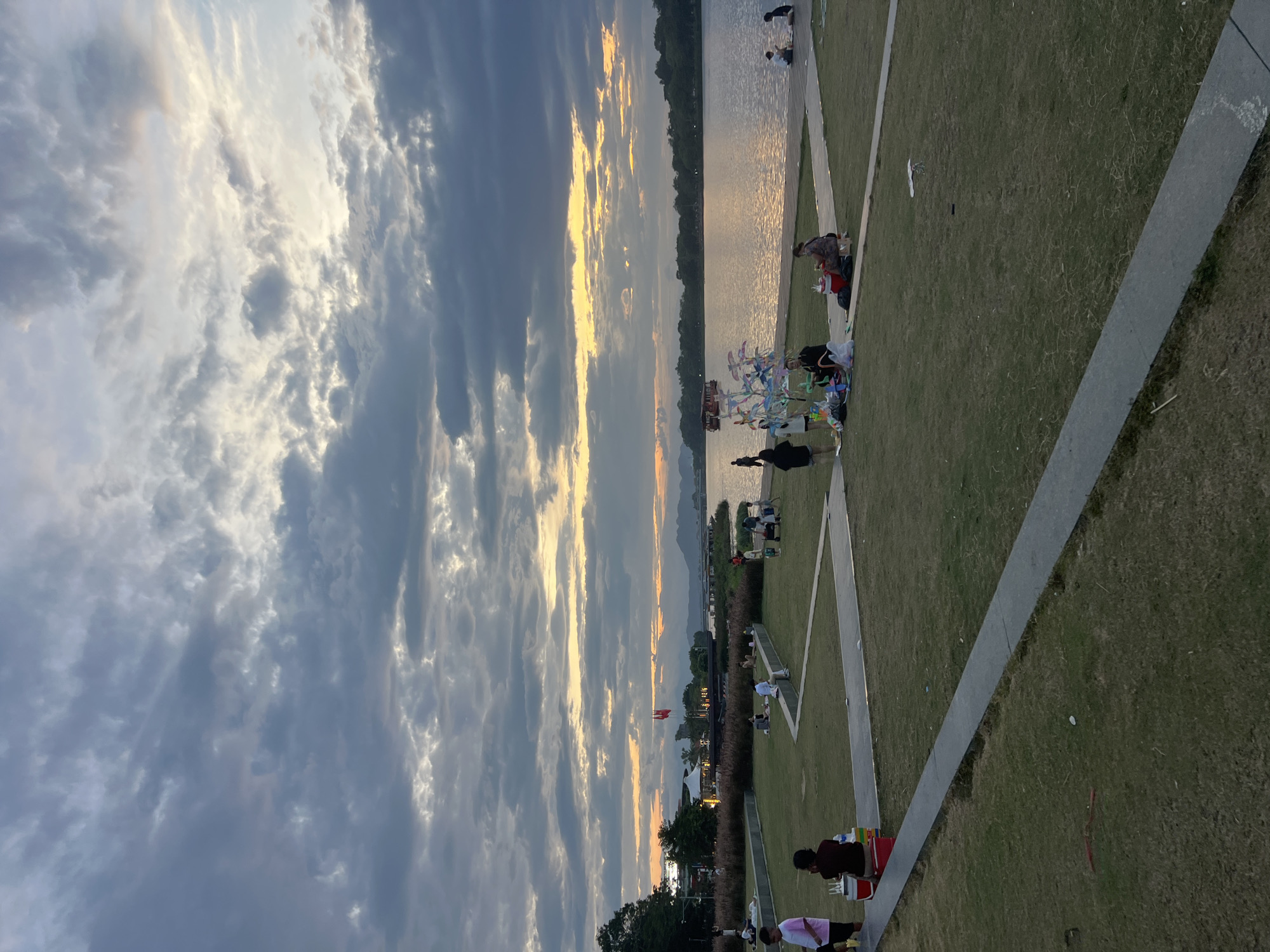The image captures a twilight scene at a waterfront park in Thuận Hóa Ward, Vietnam. The sky is dominated by dramatic, grey, and white clouds with streaks of orange and yellow near the horizon, indicating sunset. A wide body of water stretches across the mid-ground, reflecting the sky's colors.
In the foreground, a grassy park area is dotted with people. Several individuals are seated on the grass, some in small groups, appearing to be relaxing or picnicking. To the left, two children are visible near a red cooler. A decorative, colorful structure resembling a pinwheel tree stands on the grass, surrounded by a few people. Paved pathways wind through the green space.
In the middle ground, across the water, a tree-lined shore is visible, with buildings and structures, including two red flags, situated along it. A boat is moving on the water, and further in the distance, hills or mountains form a silhouette against the fading light. People are also seen scattered along the water's edge in the background on the right side of the image. The overall atmosphere suggests a calm, recreational evening by the water.