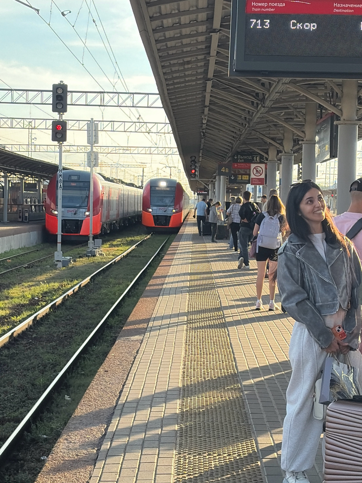 The image depicts a train station platform in Nizhny Novgorod, Russia, bathed in the warm light of late afternoon. Two red and black trains are visible, one further down the track and another closer to the foreground. A woman, the focal point of the image, stands on the platform, looking upwards and to the right with a slight smile. She is wearing a gray jacket, light-colored sweatpants, and sneakers. She is holding a small bag and a phone with a colorful case. Other people are scattered on the platform, some standing with luggage, suggesting they are waiting for trains. In the background, a digital display board shows train information, with "Train number 713" and "Скор" (meaning "Fast") clearly visible. The platform is paved with interlocking bricks, and overhead structures provide shade. Traffic signals are also present, one showing a red light. The environment is that of a functional railway station, likely a busy transit point within the city.