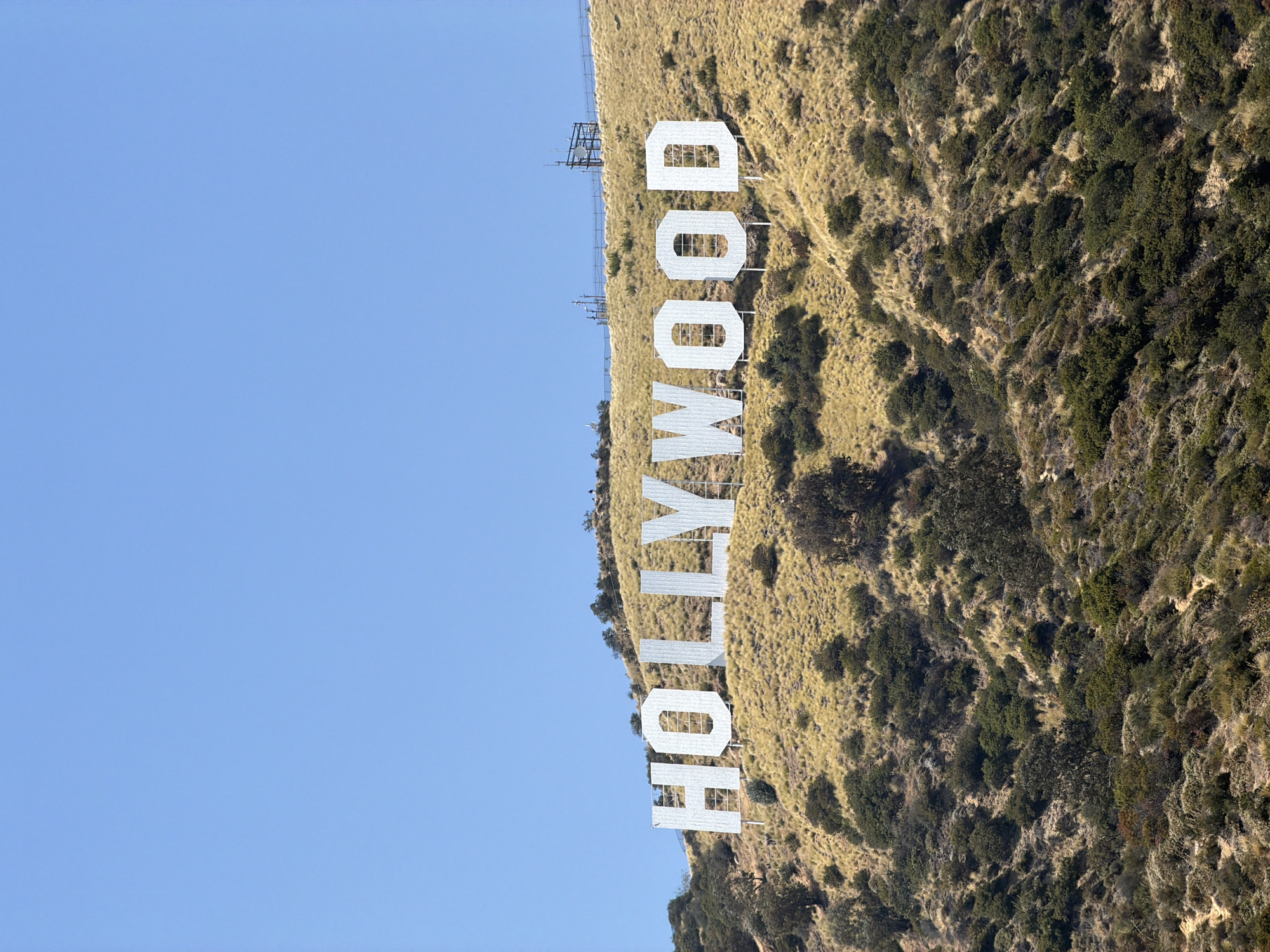 The image shows the Hollywood sign on the side of Mount Lee in the Hollywood Hills area of Los Angeles, California. The sign, spelled out in large white letters, is situated on a grassy hillside with some trees. The background is a clear, blue sky. The sign is viewed from a low angle.