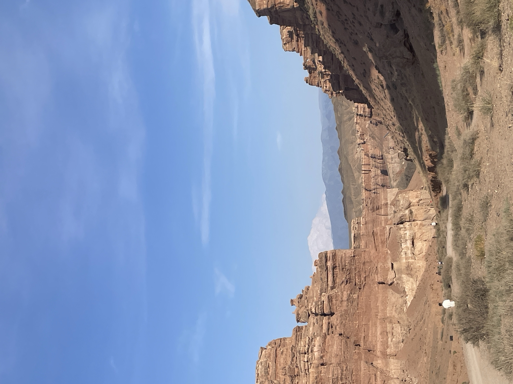 This is a photograph of the Charyn Canyon in the Tamerlik region of Kazakhstan. The image depicts a vast, arid landscape dominated by towering, eroded sandstone cliffs in hues of red and orange. The canyon floor is a dusty, uneven terrain dotted with sparse, dry brush and a dirt path winding through it. Several individuals, appearing as small figures in the distance, are walking along the path, suggesting a tourist or hiking activity. In the far background, distant mountain ranges are visible under a clear, bright blue sky with a few wispy clouds. The sunlight casts shadows that accentuate the rugged textures and depth of the canyon walls.