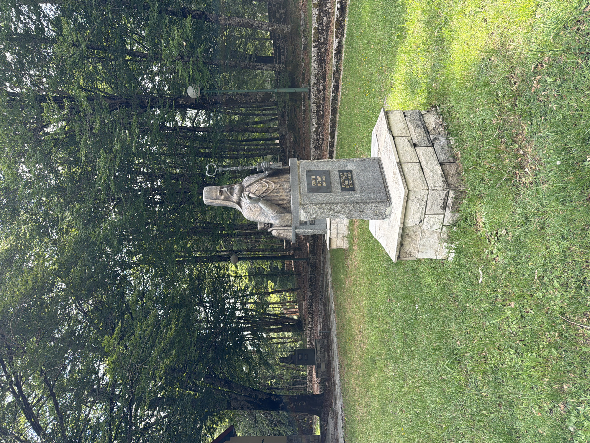 An outdoor image, rotated ninety degrees, depicts a stone monument of a historical figure set against a backdrop of dense forest and green grass. The central subject is a grey stone statue of an elderly, bearded man, likely a cleric or saint, wearing a long robe and a distinctive head covering resembling a miter. He holds a crosier (pastoral staff) in his right hand.
The statue is mounted on a multi-tiered pedestal made of lighter-colored, rough-hewn stone blocks. A dark grey plaque on the pedestal bears inscriptions in Cyrillic. The top line reads "СВЕТИ ПЕТАР" (Sveti Petar), identifying the figure as Saint Peter. Below this are the dates "1748-1830," indicating his birth and death years. Further down, the text reads "ГОСПОДАР ЦРНЕ ГОРЕ" (Gospodar Crne Gore), meaning "Ruler of Montenegro," followed by the dates "1782-1830," signifying his period of rule. This identifies the monument as commemorating Saint Peter of Cetinje (Petar I Petrović-Njegoš).
The setting is a natural environment, characterized by a thick forest of tall, leafy trees providing a shaded backdrop. A narrow, unpaved path or walkway winds through the trees in the middle distance. The ground immediately around the monument and extending into the foreground is covered in vibrant green grass. A lamp post is visible within the forest to the left of the statue. Another smaller, darker object, possibly a sign, can be seen along the path further back. The lighting suggests it is daytime, with natural light filtering through the trees. No specific activity is occurring. The location is Dolovi, Montenegro.