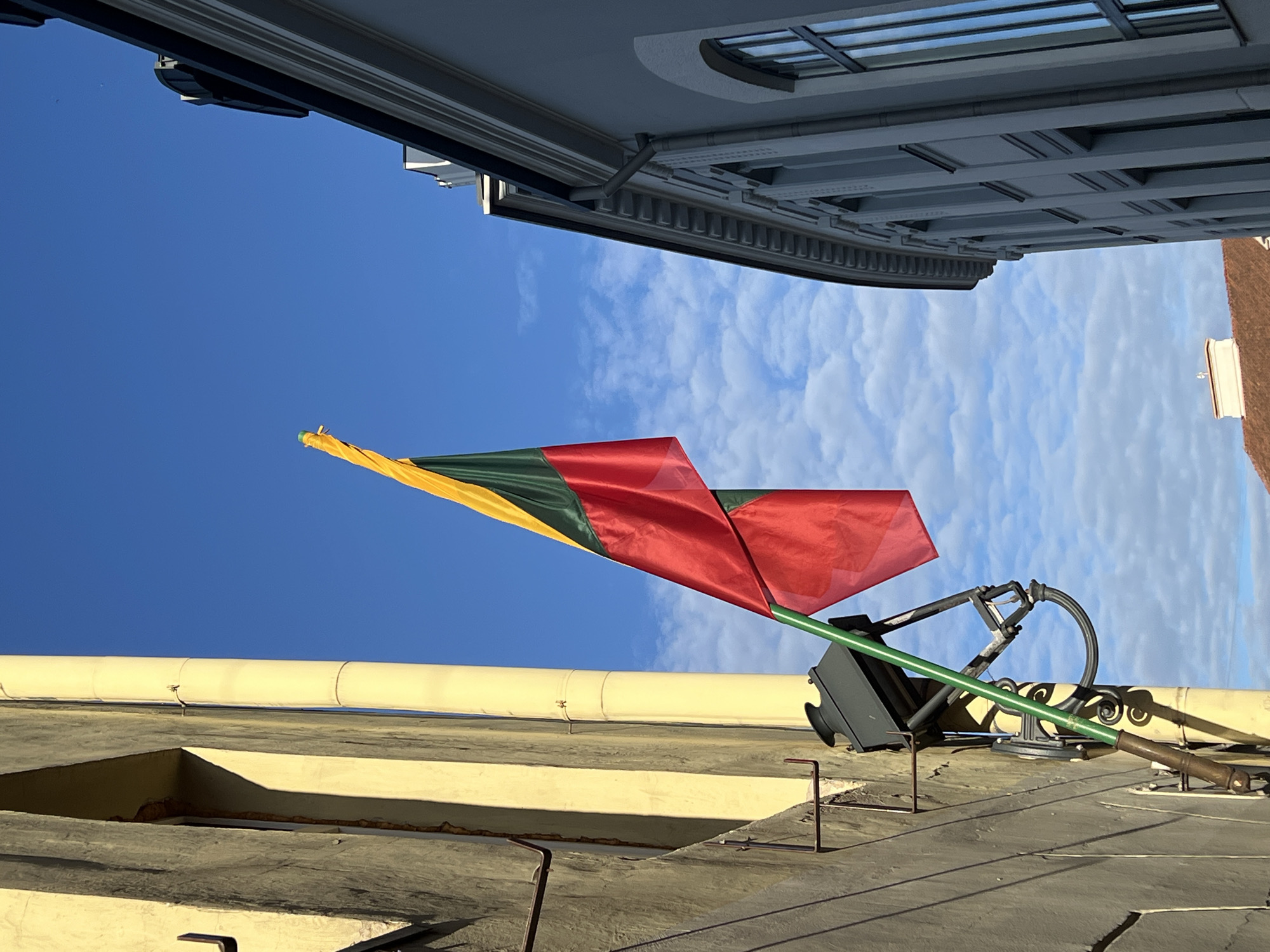 The image captures a Lithuanian flag, composed of horizontal stripes of yellow, green, and red, suspended against a bright blue sky with scattered clouds. The flag is mounted on a green pole, attached to the side of a building with a pale yellow facade. The building has a textured surface and features a dark, old-fashioned street lamp fixture mounted near the base of the flag pole. To the right of the flag pole, a portion of another building with a light grey facade is visible, characterized by its ornate architectural details and multiple windows. The perspective suggests the photo was taken from a low angle, looking upwards between two buildings in what appears to be a narrow street or alleyway. The setting is urban, consistent with the historic architecture found in Vilnius, Lithuania. There are no people visible in the image.