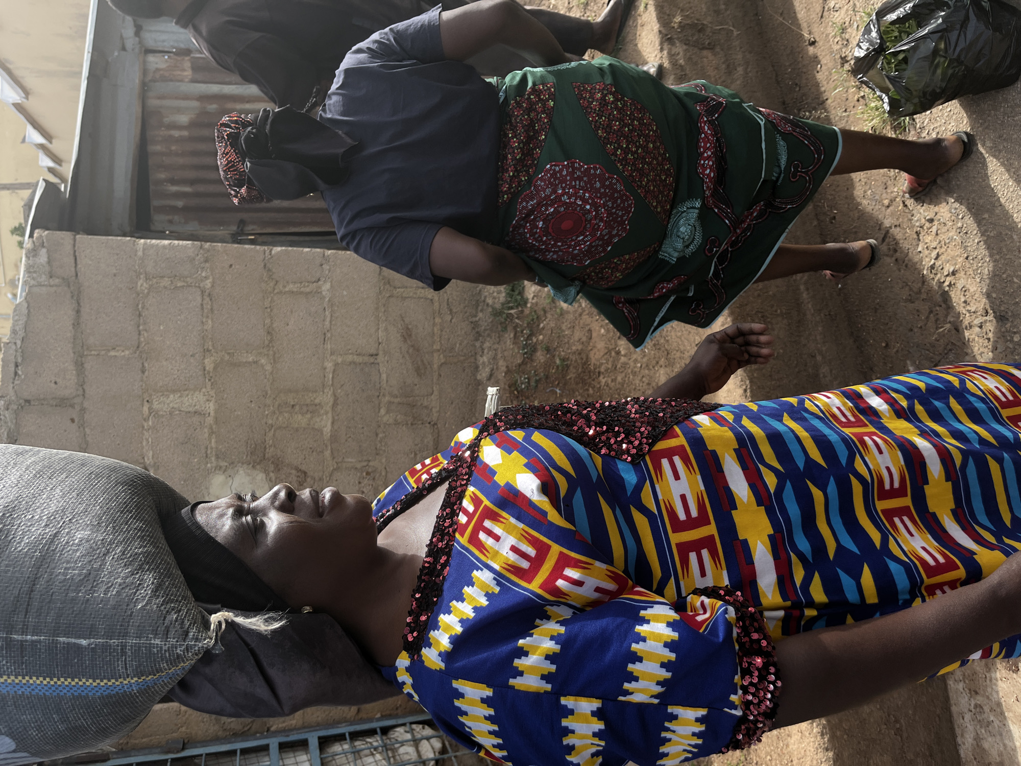 A medium shot, taken outdoors, shows two women in Jos, Nigeria. The woman in the foreground faces left with a large grey bag balanced on her head. She wears a blue dress patterned with yellow, red, and white geometric designs, and a sequined collar. The second woman, further back, wears a dark blue shirt and a green skirt with red circular patterns. She is also carrying a black plastic bag. The background includes a brick wall and a corrugated metal structure. The ground is sandy and uneven.