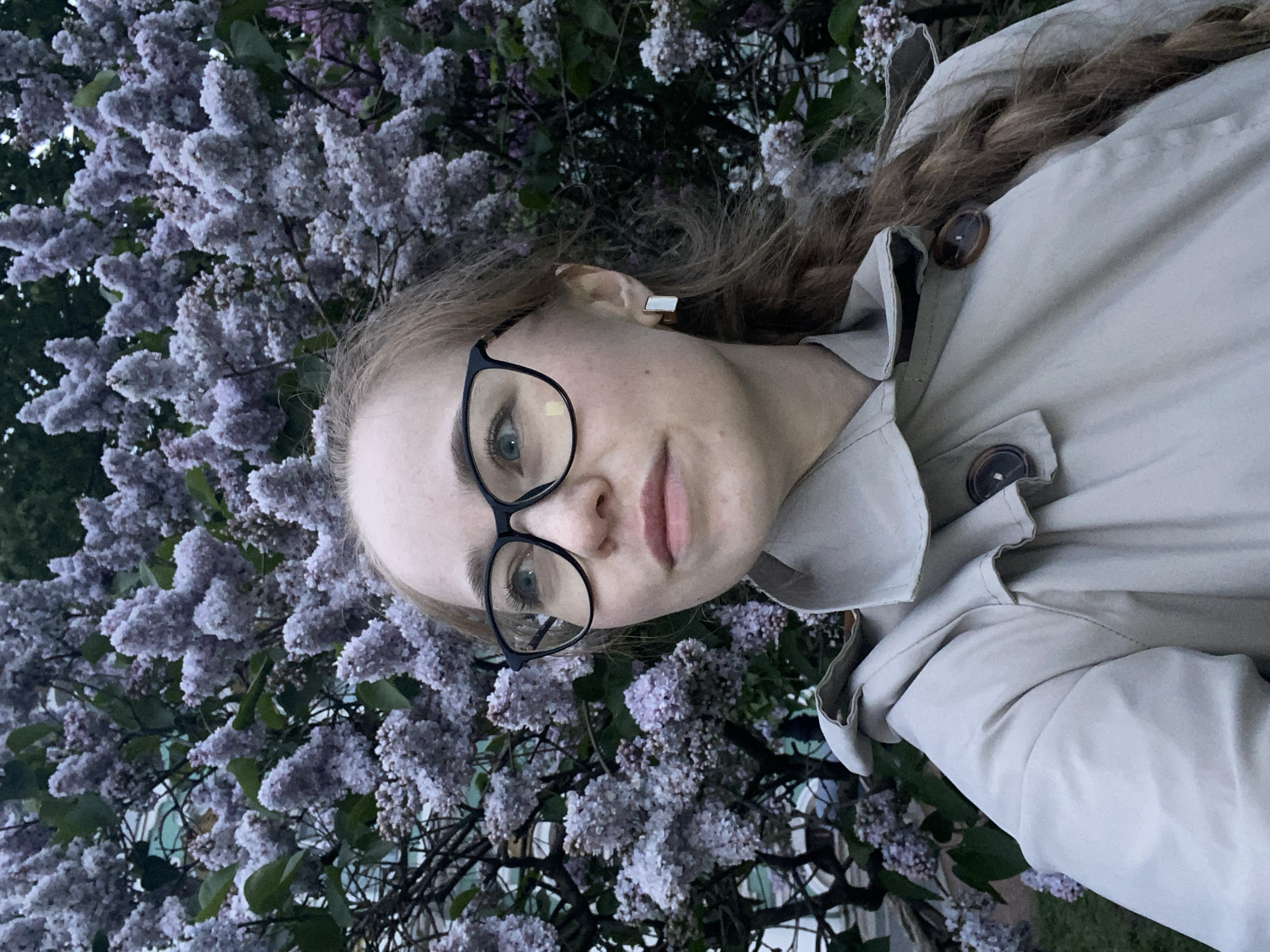 A young woman with blonde hair, wearing glasses and a tan trench coat, stands in front of a lilac bush. The woman is facing the camera and slightly tilted to the side. The lilac bush is full of purple flowers. The location is likely Saint Petersburg, Russia, due to the prevalence of lilac bushes in the city.