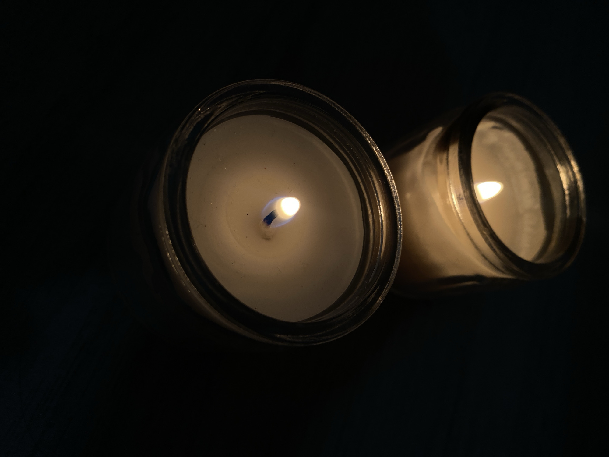 The image presents an overhead view of two lit candles in clear glass jars against a profoundly dark, unidentifiable background. The candle in the foreground, positioned prominently on the left, is sharply in focus. Its bright flame, featuring a distinct blue hue at its base, illuminates the white wax and the interior of the glass jar, casting a soft, warm glow.
To the right and slightly behind, a second candle in a similar clear glass jar is partially visible and slightly out of focus, also emitting a gentle, warm light from its flame. The surrounding environment is completely dark, suggesting either a nighttime setting or a deeply shadowed room where the candles are the sole sources of illumination. Subtle reflections are visible on the glossy surfaces of the glass jars, and some minor textural details or accumulated wax can be observed along their rims. The overall scene conveys a sense of calm and warmth, emphasizing the isolated radiance of the flickering flames in the darkness. No text is visible in the image.