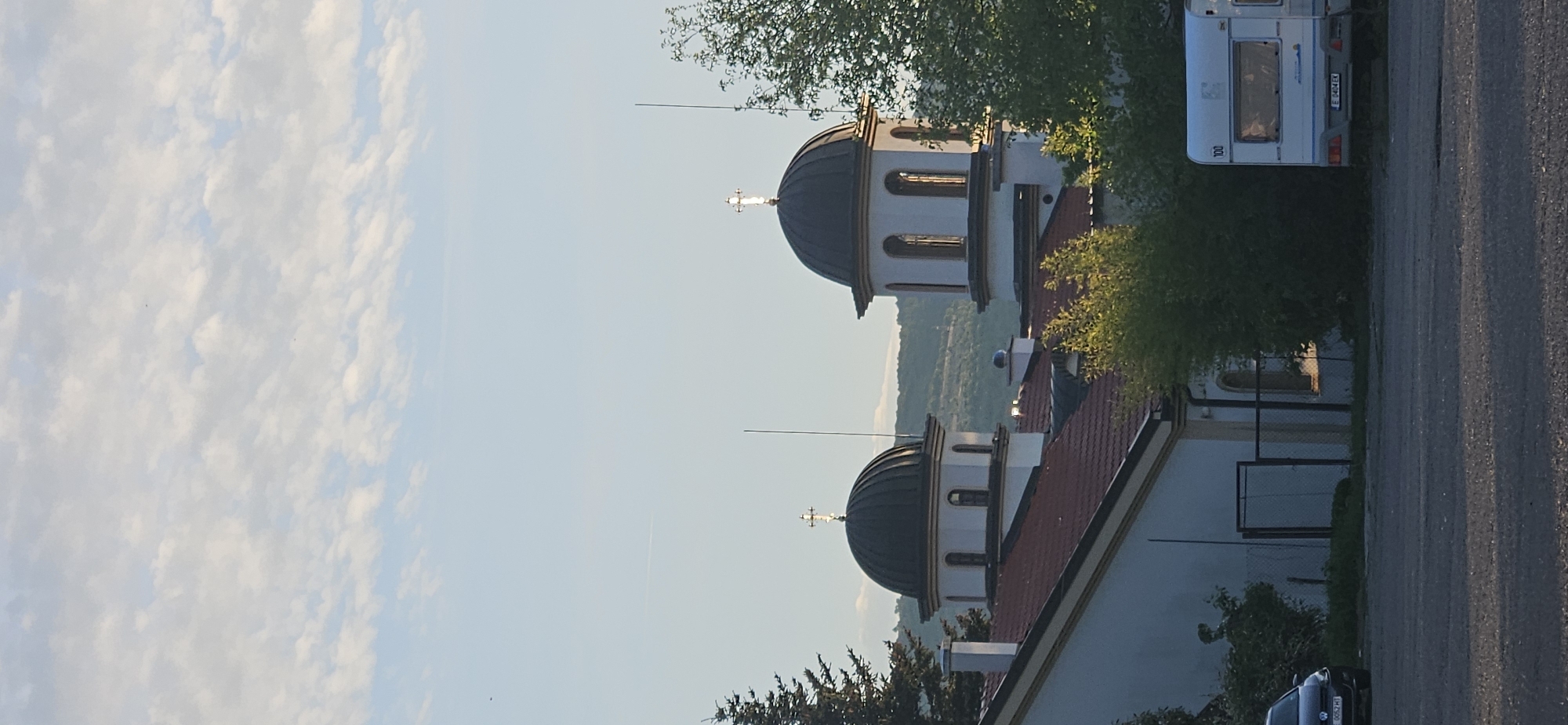 The image shows an Eastern Orthodox church located in Blagoevgrad, Bulgaria. The church has two onion domes, topped with crosses. The church is white with red trim. Behind the church, there is a hill covered with trees. In the background, there is a blue sky with clouds. A camper van and a car are parked on the side of the church.