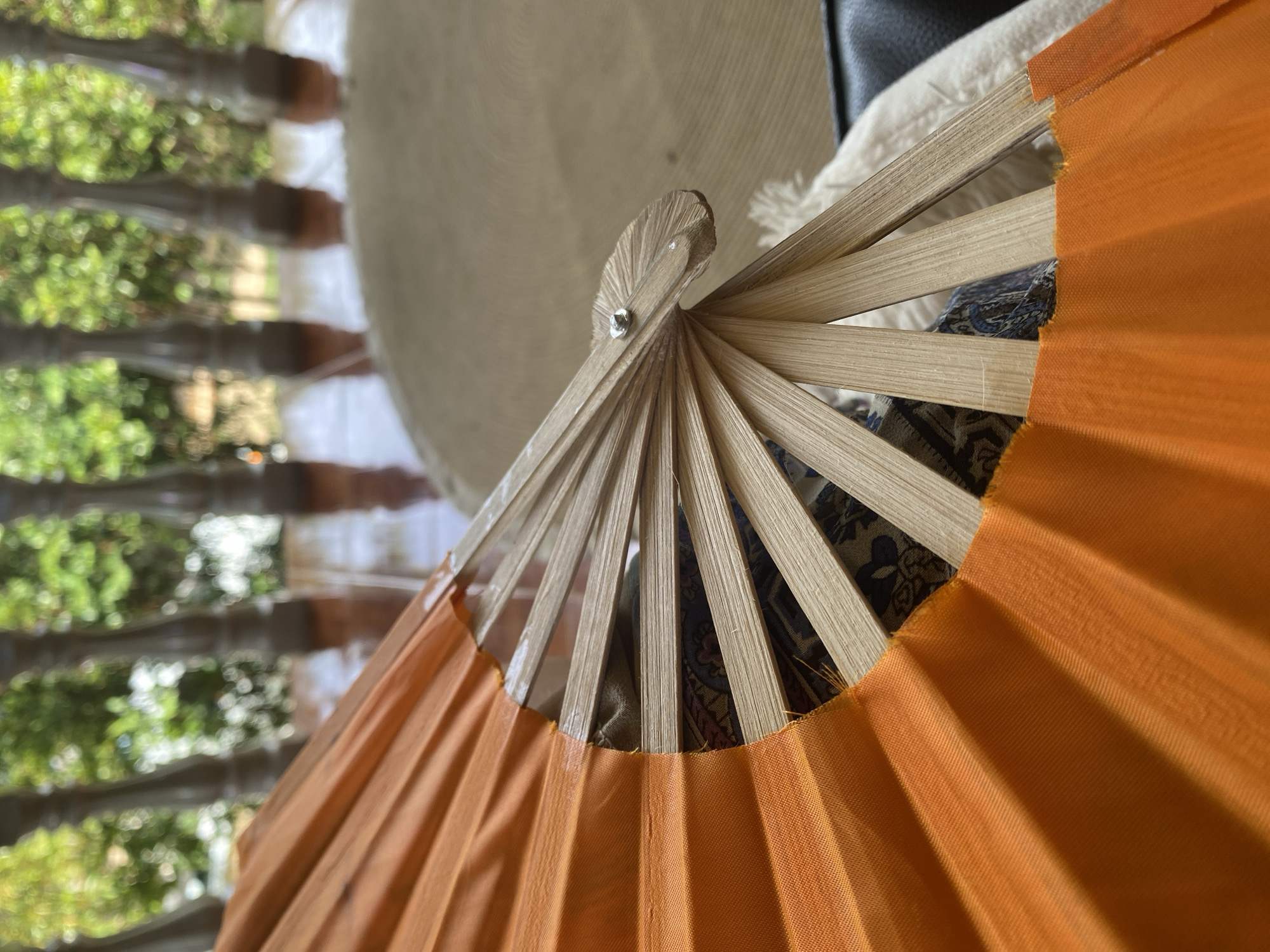 A close-up shot depicts an open hand fan with orange fabric and light-colored wooden slats. The central pivot point of the fan is visible, held together by a small metal fastener. The fan partially obscures a background with blurred elements including what appears to be foliage seen through blinds. The location is Baan Nai Suan, Thailand.