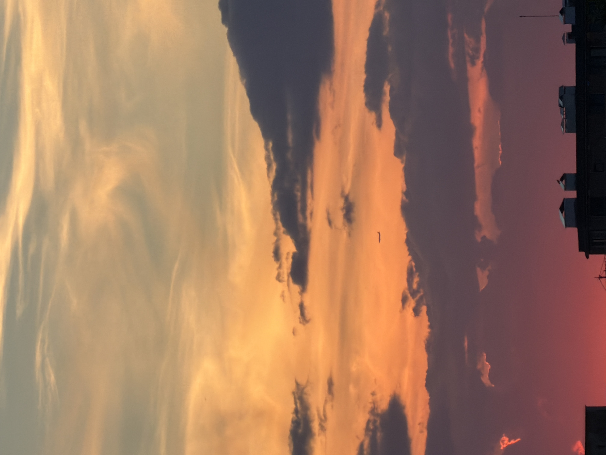 The image depicts a dramatic sunset over an urban environment, likely Sofia, Bulgaria. The sky is filled with a breathtaking display of clouds painted in vibrant hues of orange, pink, and deep purple. Wispy, cirrus clouds catch the last rays of sunlight, creating streaks of gold against the darkening sky. Darker, more turbulent clouds are also present, adding depth and contrast to the scene.
In the background, the silhouette of buildings can be seen, suggesting a city skyline. The buildings are dark and indistinct, their forms softened by the fading light. One building on the right has several visible air conditioning units or similar rooftop structures. A single, dark bird is visible in flight against the colorful sky, adding a dynamic element to the composition.
The time of day is clearly sunset, with the intense colors of the sky indicating the sun has recently dipped below the horizon. The weather appears clear enough for a spectacular sunset, with no indication of rain or storms. The overall mood is one of awe and natural beauty, with the urban landscape serving as a backdrop to the celestial spectacle. There is no visible text in the image.