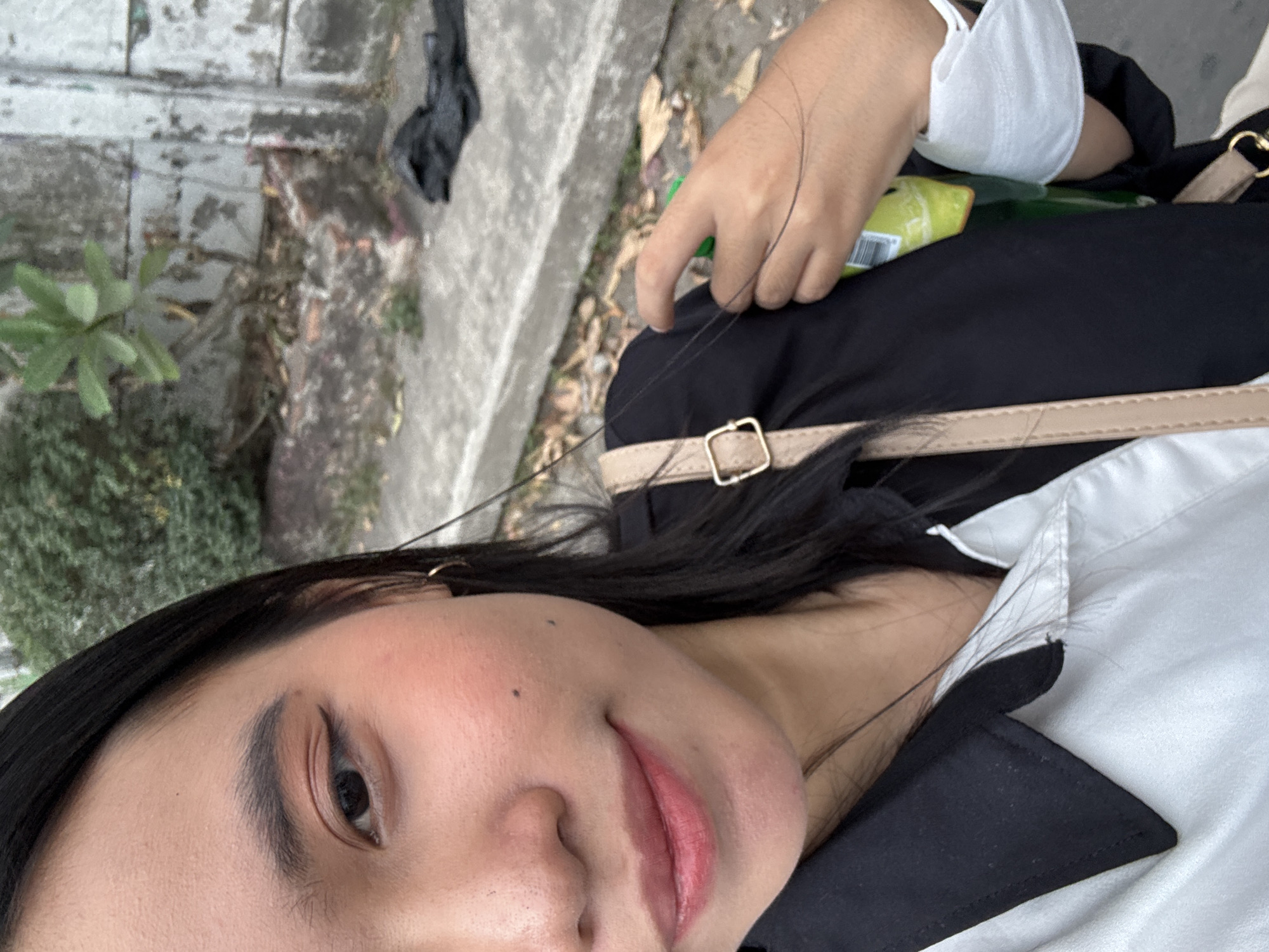 A close-up, waist-up, selfie-style photograph features a young woman with black hair and fair skin. She is wearing a white collared shirt with black accents on the collar and sleeves, and a black outer garment. A beige crossbody bag with a gold buckle is visible, slung across her body. She has a light makeup application, including eyeliner and blush. In her right hand, she is holding a green bottle and a white face mask. The background suggests an outdoor urban setting with a grey concrete wall, pavement, and some vegetation. The image is taken during the daytime.