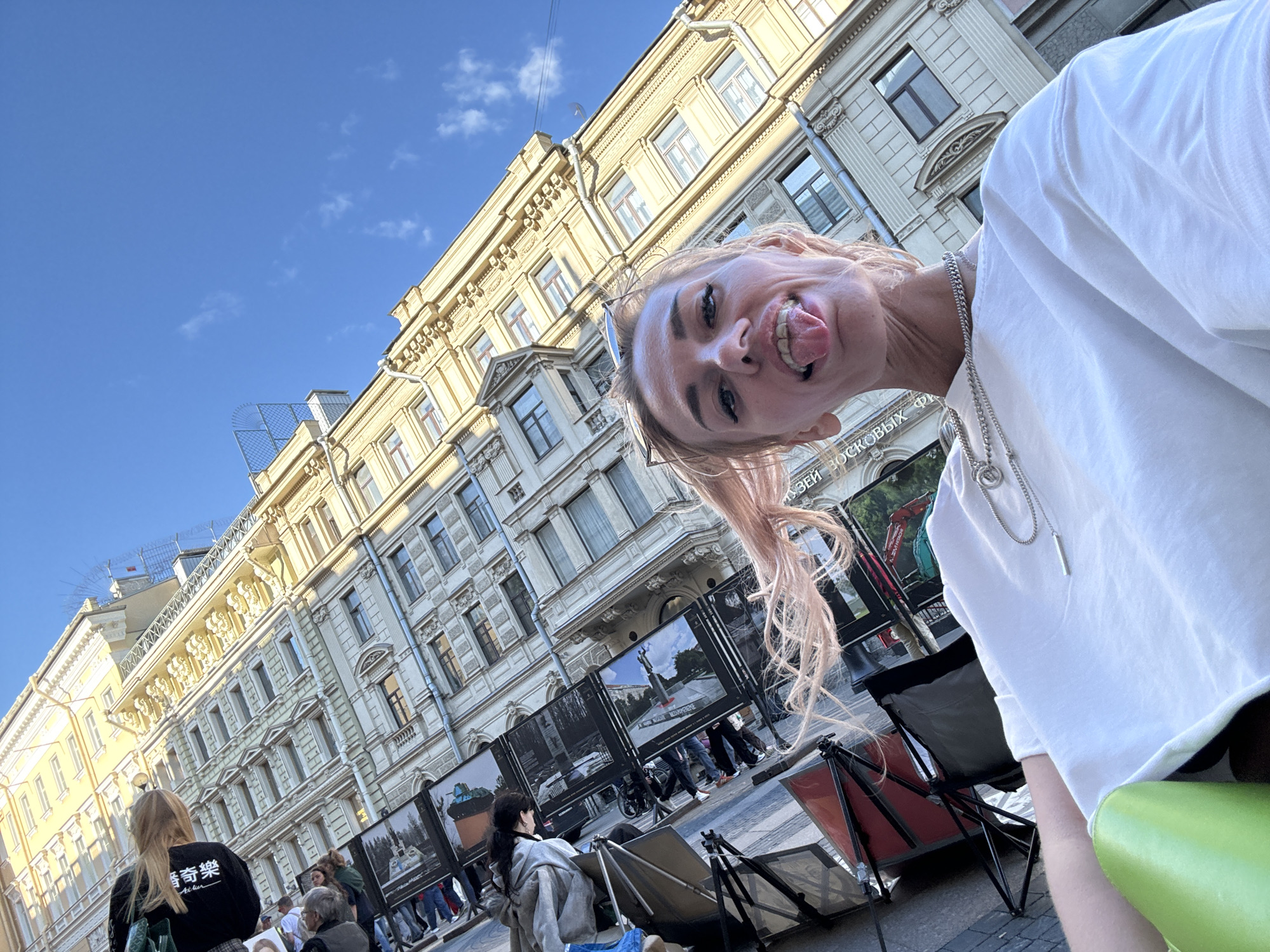 This is a selfie taken in Saint Petersburg, Russia, on a sunny day. A young woman with blonde hair is sticking her tongue out at the camera, leaning into the frame from the right. She is wearing a white t-shirt and a silver necklace. In the background, a light-colored building with ornate architectural details lines a city street. Several framed photographs are displayed on easels on the sidewalk, suggesting an outdoor exhibition. Other people are visible in the background, looking at the displays or walking by. The sky is bright blue with a few wispy clouds.