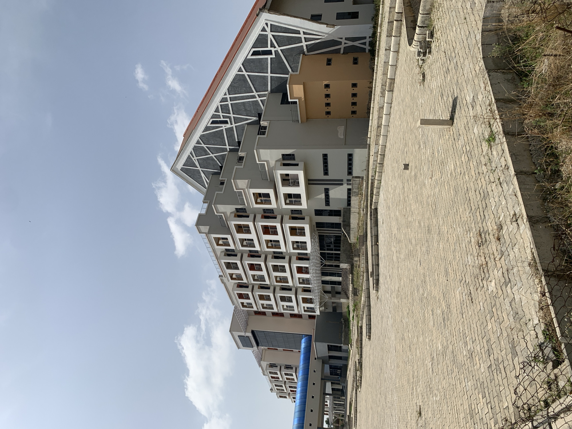 The image shows a modern, multi-story building in Jos, Nigeria. The building is primarily gray with a striking, geometric design on its roof. Below the roof, there are sections of the building with rectangular windows and balconies. A blue, covered walkway connects two sections of the building at the lower levels. In the foreground, a paved walkway leads towards the building, bordering an area of dry vegetation and a wire fence. The sky is light blue with scattered clouds. There are no people visible in the image.