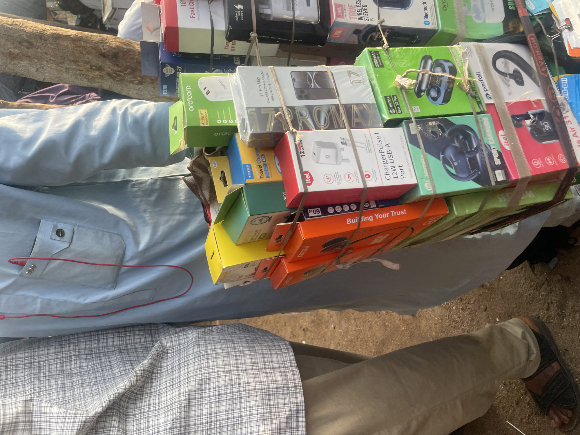 In Wauro Jabbe, Nigeria, a vendor displays a stack of electronic accessories for sale. The vendor, wearing a blue traditional outfit with a red earpiece plugged into a pocket, stands next to a person in a checkered shirt and beige trousers. The accessories, primarily chargers and earbuds, are neatly packaged in colorful boxes. They are arranged in a tiered display, bound together with twine. In the background, a tree trunk and other indistinct figures and objects suggest a bustling marketplace environment. The ground beneath the display is sandy.