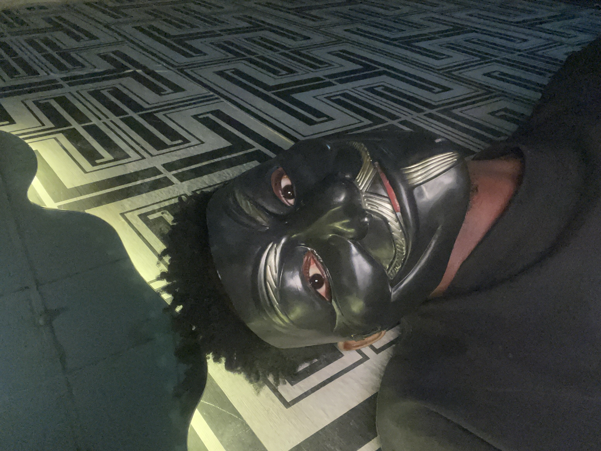 A person wearing a Black Panther mask lies on a patterned floor. The floor has a repeating geometric design in shades of grey and white. The person wears a dark colored sweatshirt. The location cannot be identified as Lokoja, Nigeria, based solely on the provided image.