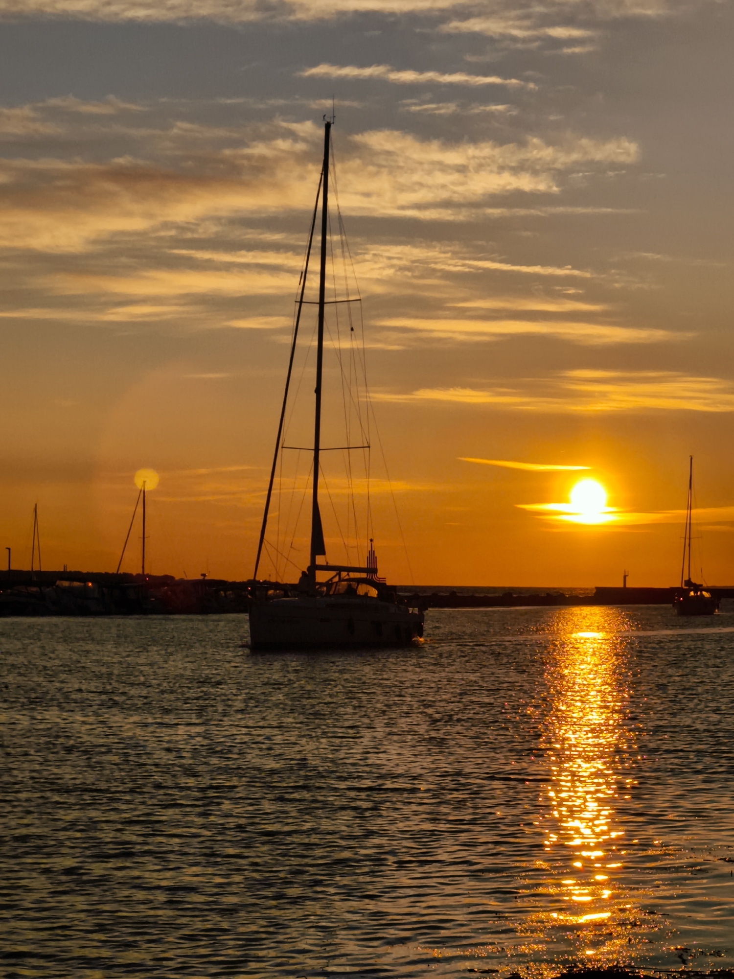 This is a photograph of a sunset over a body of water, likely a harbor or bay, with several sailboats visible. The sun is low on the horizon, casting a warm, golden glow across the sky and reflecting on the water's surface. The sky is filled with scattered clouds, tinged with orange and yellow. The foreground shows the rippling water, with a prominent reflection of the setting sun. Silhouettes of boats and a distant pier are visible in the midground and background. The image evokes a sense of tranquility and natural beauty. There is no indication of this specific location being in Tallinn, Estonia.