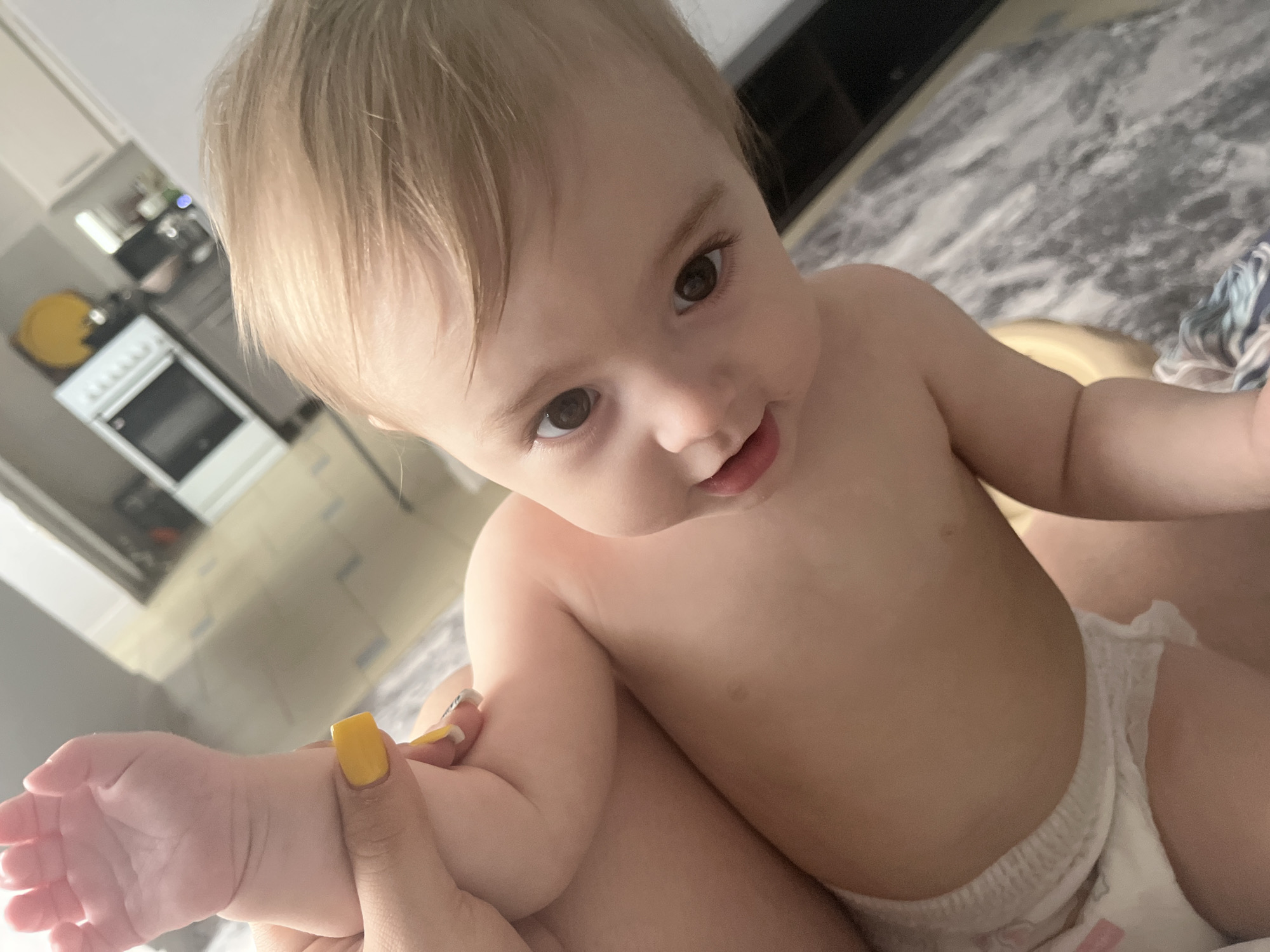 A baby is seated on a person's lap, likely indoors. The baby is fair-skinned with light brown hair, brown eyes, and is wearing a diaper. The person holding the baby has a fair skin tone and yellow nail polish. The background shows a kitchen with white cabinets and appliances, including an oven. The floor appears to have a gray and white pattern. The location is unspecified, with no specific indicators relating to Chaikovsky, Russia.
