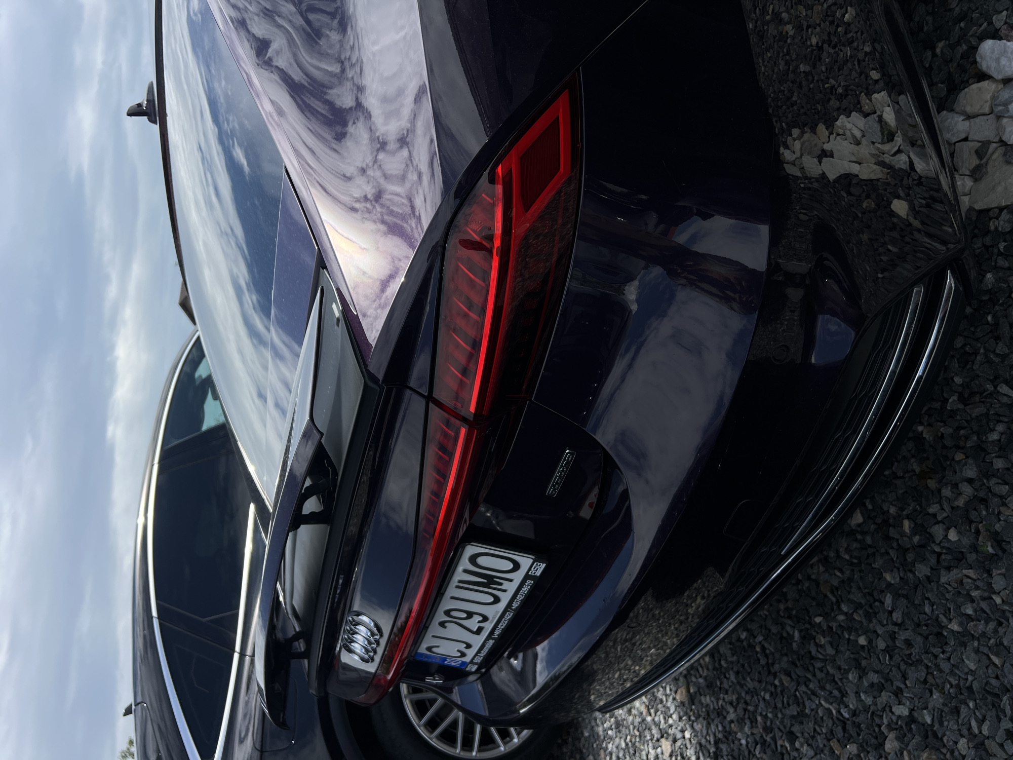 This media file shows the rear of a dark-colored Audi. The car is parked outdoors on a surface of dark grey and white gravel. In the background, part of another car is visible, along with a cloudy sky. The car's taillight is illuminated red, and the license plate is clearly visible.
The license plate reads "CJ 29 UMO" and displays several lines of smaller text below the main characters. Some of this text includes "RSB Automobile", "RO", and a series of numbers that appear to be a VIN or identification code. The Audi logo, a series of four interlocking rings, is visible on the left side of the rear of the car, near the taillight. There is also an emblem on the right side of the rear, below the taillight, which appears to say "quattro".
The image is taken from a low angle, looking up at the rear of the car. The lighting suggests it is daytime, and the cloudy sky indicates overcast weather. There are no people visible in the image. The scene appears to be a parking area or lot, possibly in Cluj-Napoca, Romania, given the "CJ" on the license plate, which is the abbreviation for Cluj County. The overall impression is of a static, detailed view of a parked luxury vehicle.