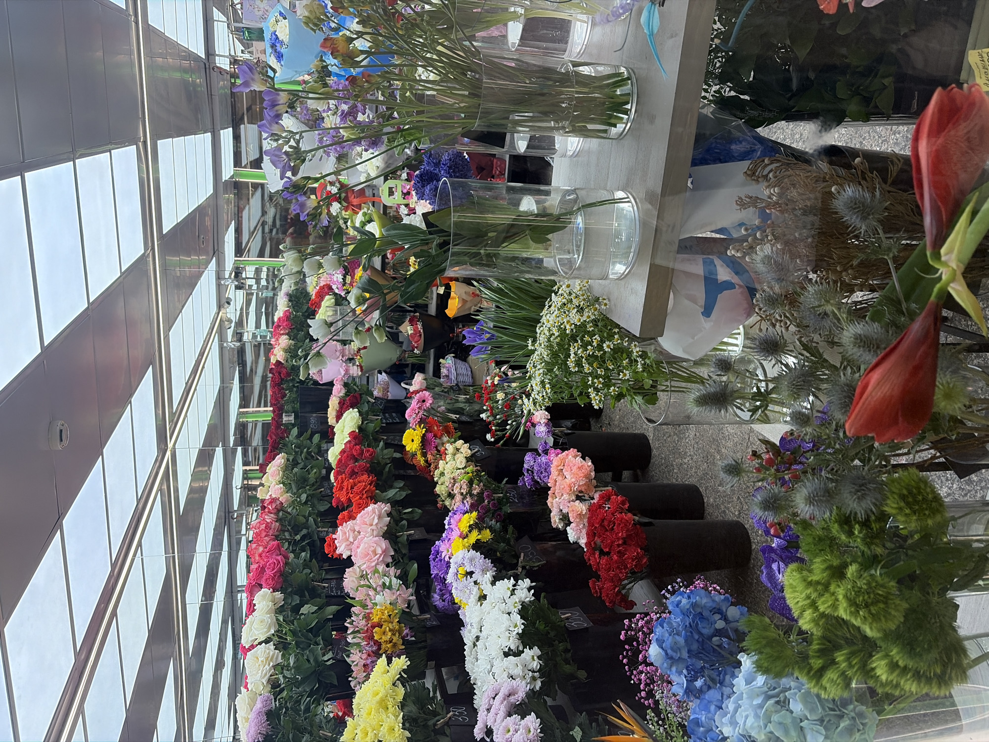 The image depicts a floral display in what appears to be a florist shop or market stall. Rows of flowers in various colors (yellow, white, pink, red, purple, blue, etc.) are arranged in black containers. Vases of cut flowers are arranged behind the rows. The location may be a shop or market in Saint Petersburg, Russia, although this cannot be determined conclusively from the image.