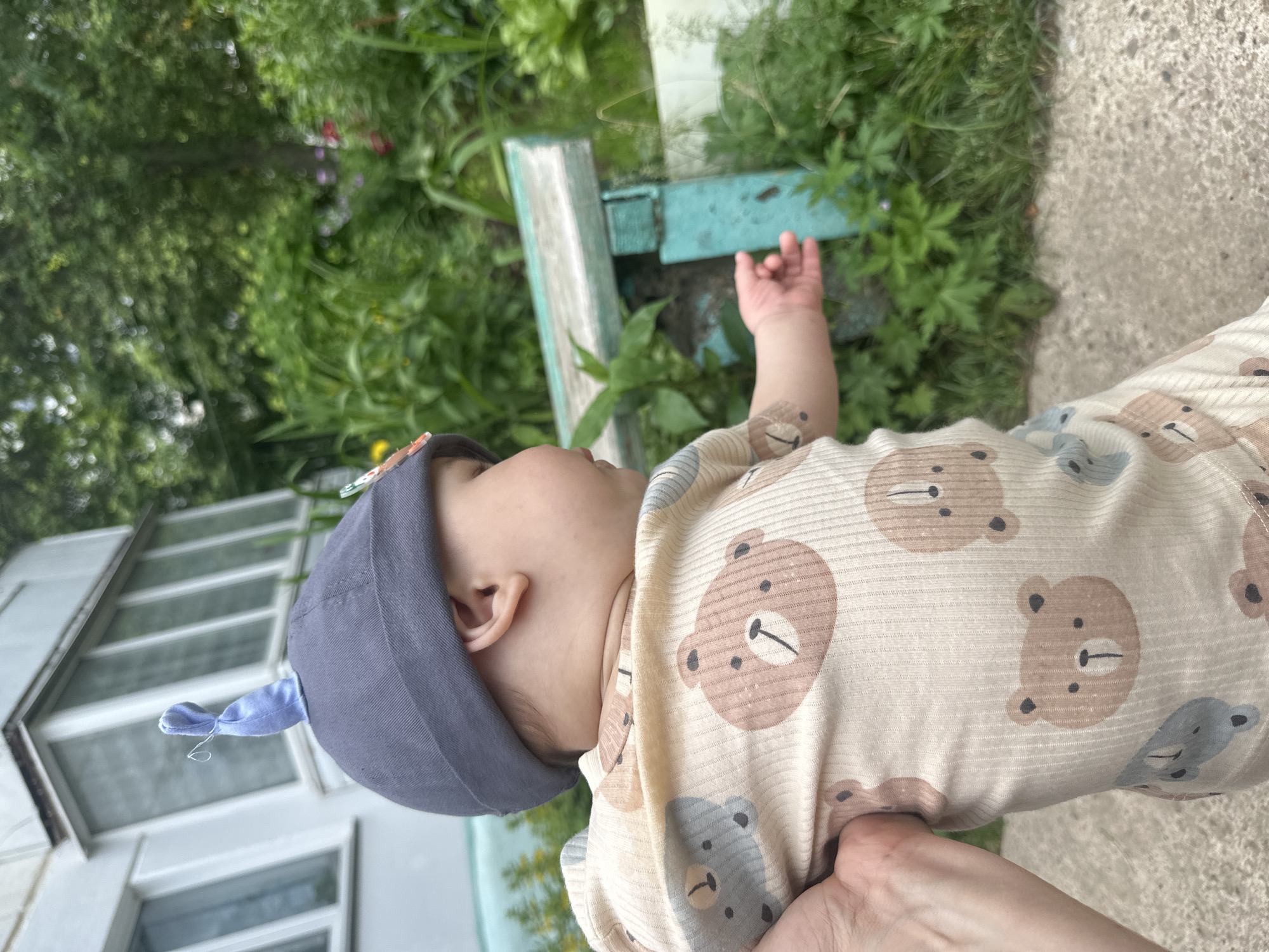 In this medium shot, a baby wearing a grey hat and a onesie with a bear print is held by a hand on the left of the frame, facing right. The baby's head is turned to the right, with their mouth slightly open and their gaze directed downwards. The background features lush green foliage, a turquoise bench, and a white building with a balcony. The setting appears to be outdoors, possibly a yard or garden. The image captures a candid moment of a baby being held outdoors, with a focus on the baby's profile and their interaction with the environment.
