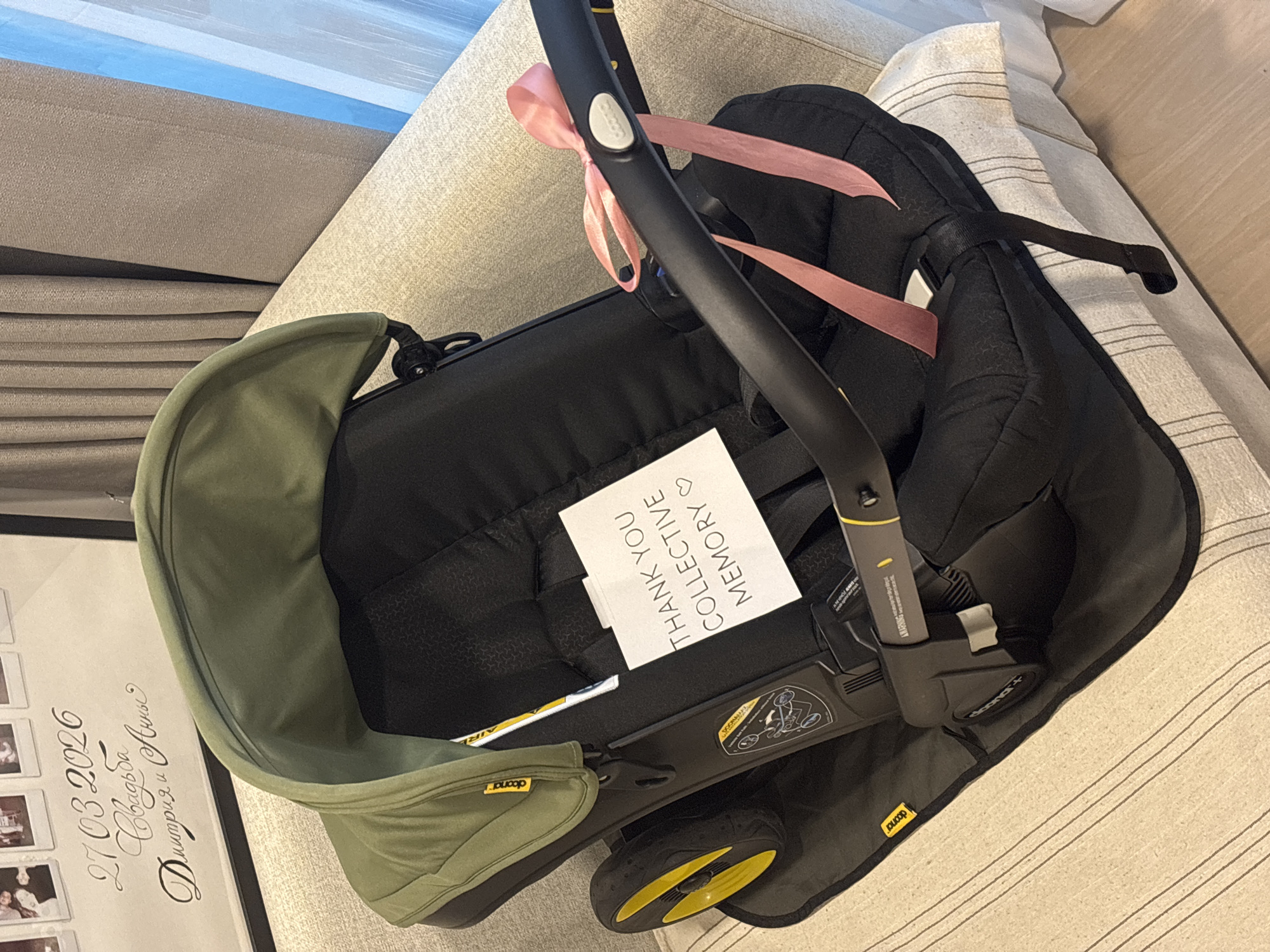 A Doona car seat/stroller hybrid is centrally positioned on a striped beige couch, indoors. The car seat is predominantly black with a green sunshade. A pink ribbon is tied around the handle. A white card with the text "THANK YOU COLLECTIVE MEMORY" is tucked inside the car seat. The location cannot be identified as being in Choeng Thale, Thailand based on the given image.